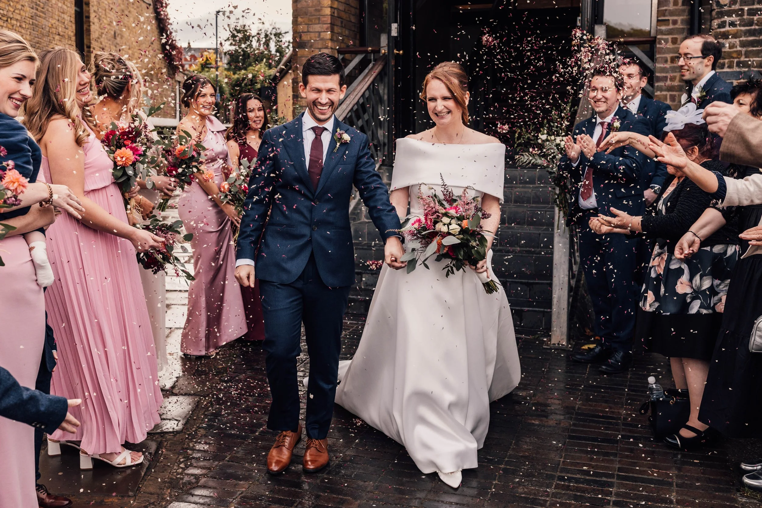 A newlywed couple holding hands and smiling while walking outside a building, with the bride carrying a bouquet of red and white flowers.