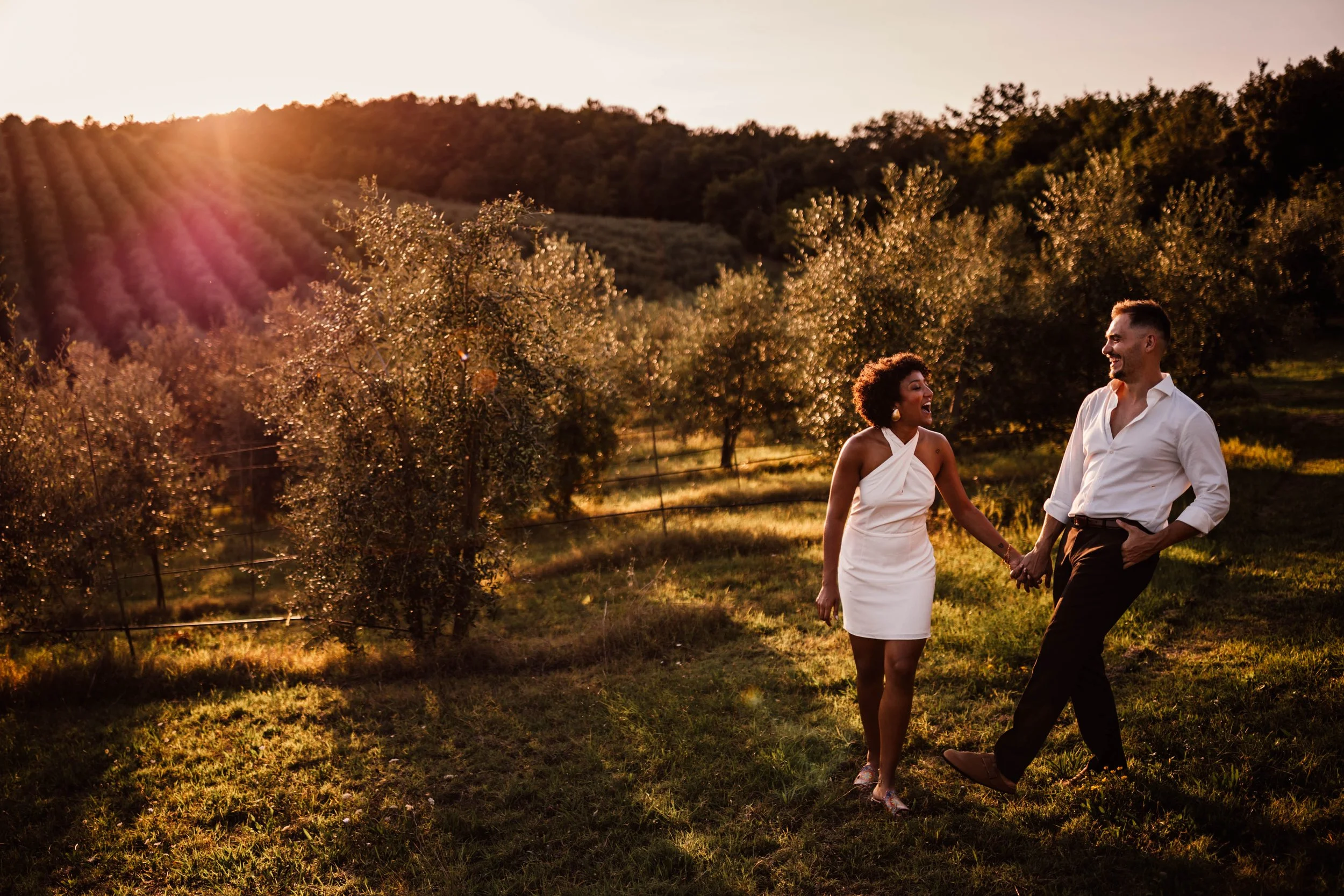 A couple holding hands and smiling while walking through a vineyard at sunset.