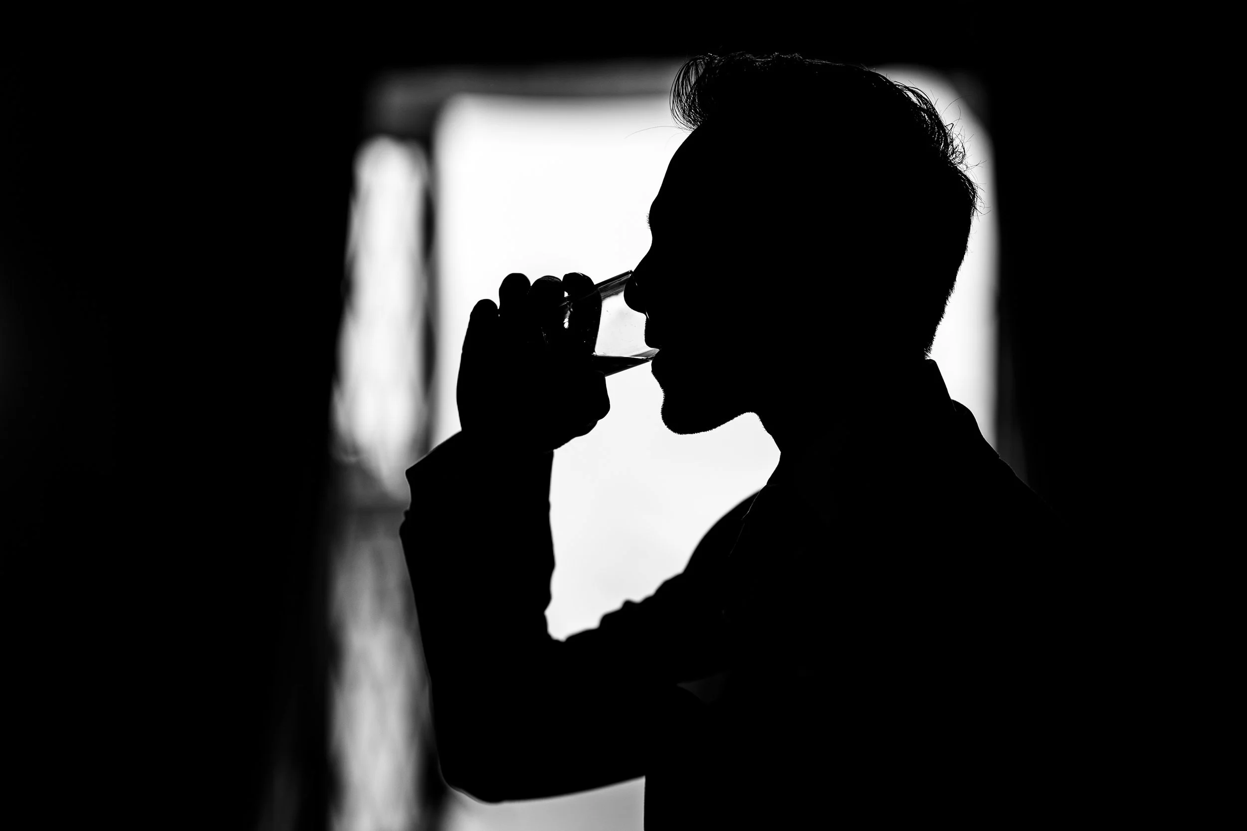 Silhouette of a man drinking from a glass, holding it near his lips, with a bright window in the background.