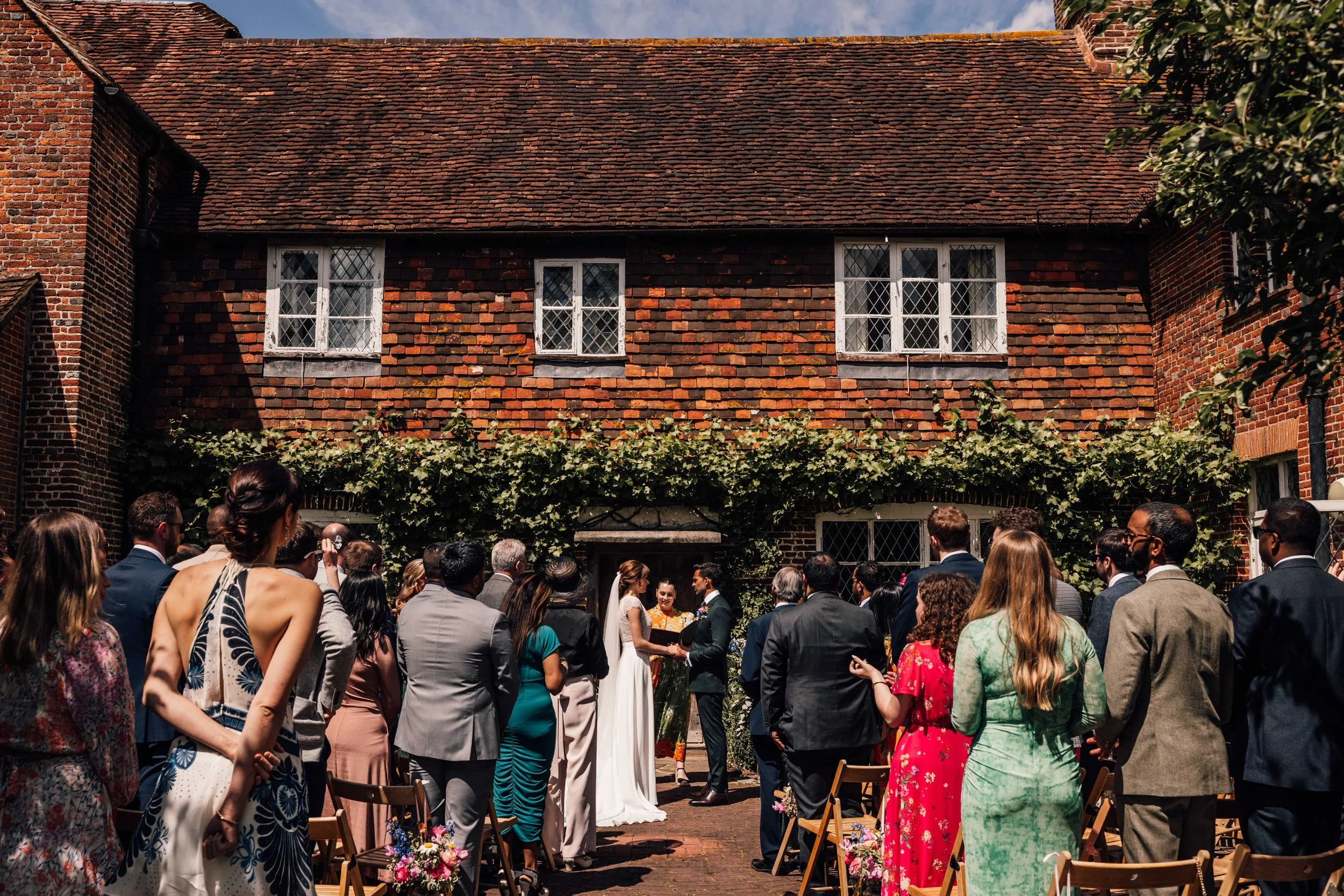 A wedding ceremony taking place outdoors in front of a brick building with ivy, with guests standing around watching a bride and groom exchange vows.