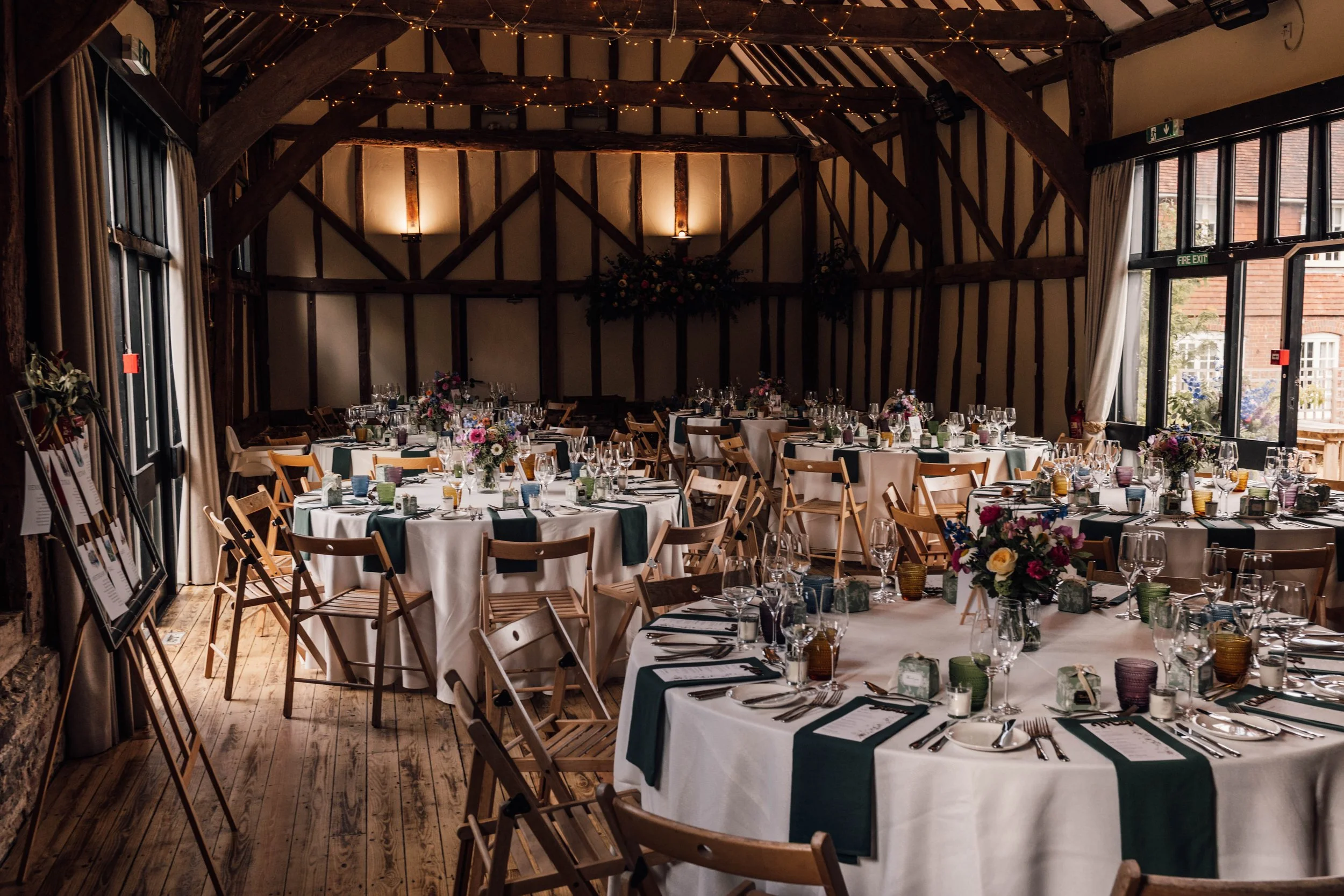 A decorated interior of a rustic wedding or event reception venue with round tables covered in white tablecloths, set with glassware, cutlery, and floral centerpieces. The venue has wooden beams, large windows with curtains, and soft lighting.