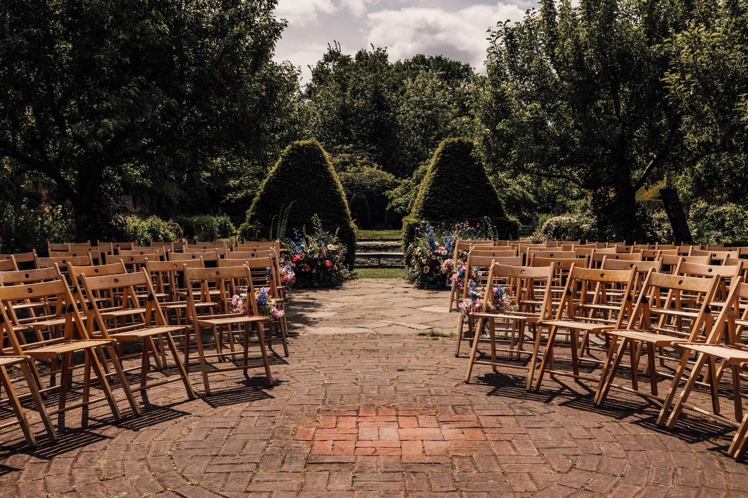 Outdoor wedding ceremony setup with wooden chairs arranged on a brick pathway, floral arrangements, and lush green trees in the background.