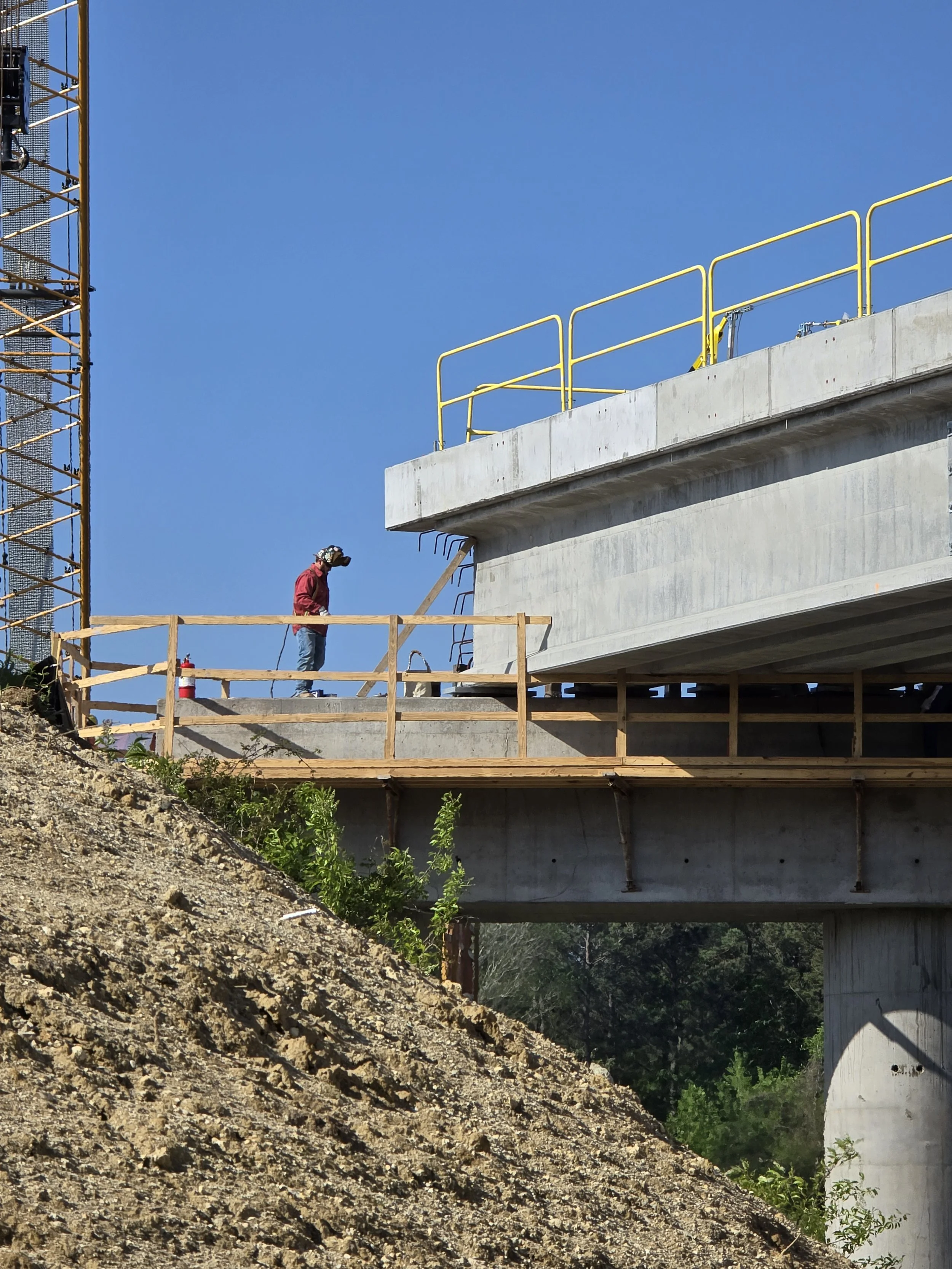 Heavy equipment positions fabricated steel beneath a concrete bridge deck as Melting Point industrial construction crew perform structural installation on a major infrastructure project for Kiewit.