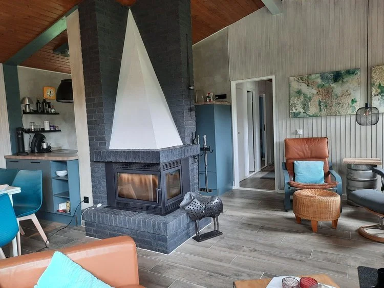 Moderner Wohnraum mit Kamin, bequemen Sesseln, Kissen, Stehlampe, Wandbildern, Küchenecke mit Kaffeemaschine, Regalen und dekorativen Gegenständen.