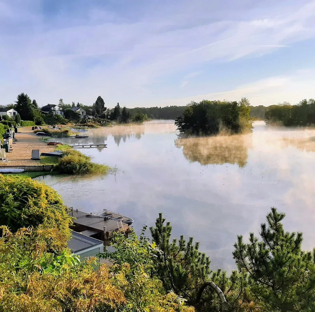 Landschaftsaufnahme des Prüßsees mit Nebel, umgeben von Häusern, Bäumen und einem grünen Ufer.