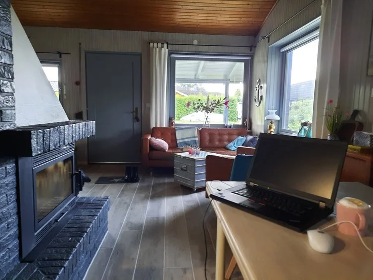 Gemütliches Wohnzimmer mit Kamin, Sofa, Laptop auf Schreibtisch, großen Fenstern mit Blick auf den Garten, Deko und Pflanzen.