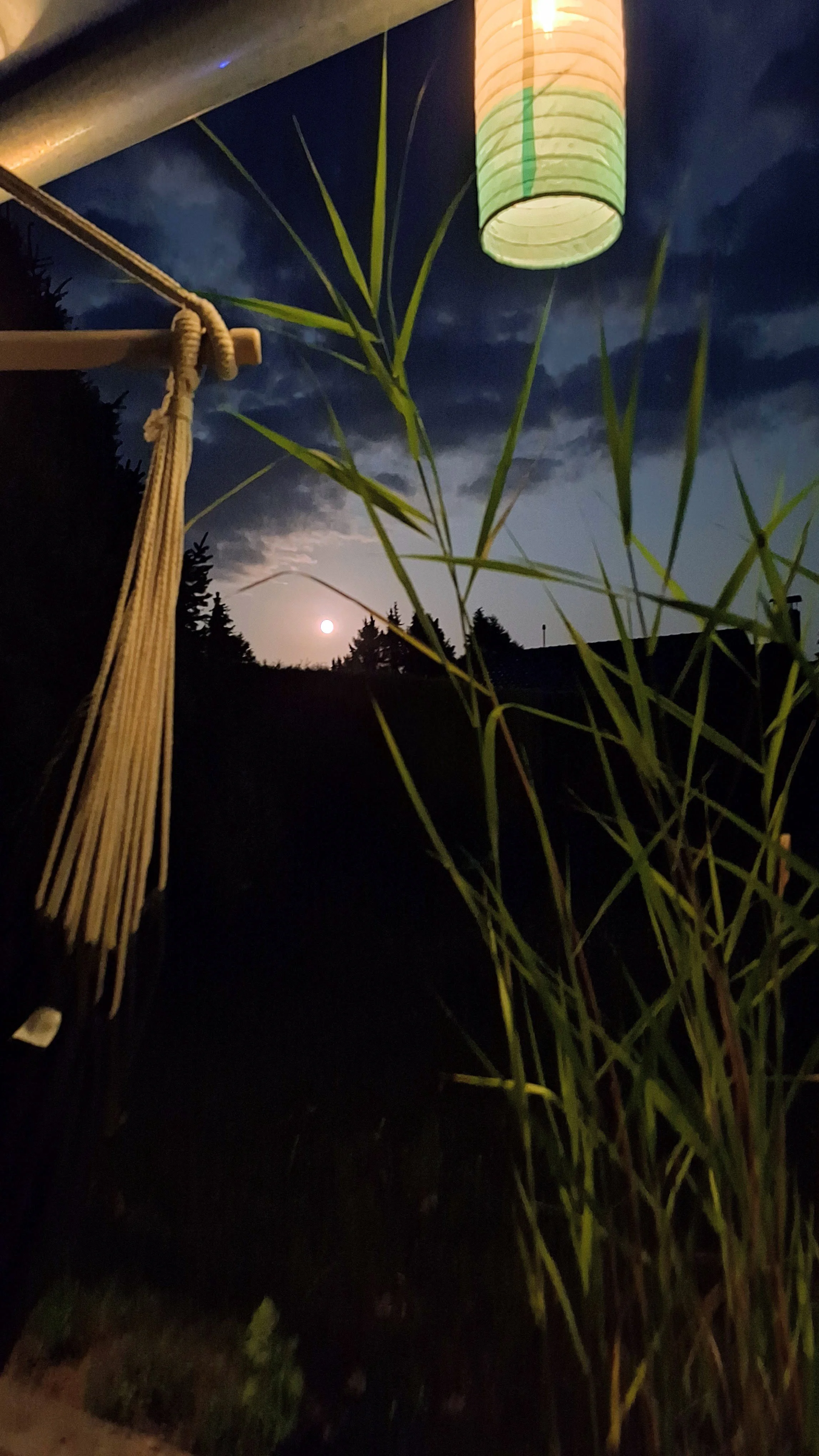 Nachtaufnahme mit Vollmond, Wolken, Gräser, einer Hängematte und einer bunten Papierlaterne im Vordergrund.