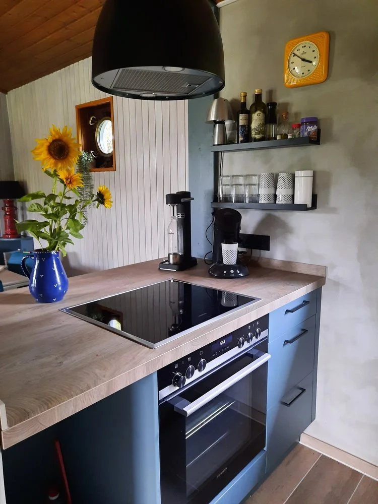 Küchenbereich mit Herd, Mikrowelle, Wasserkocher, Gläser, Sonnenblumen in blauer Vase, Uhr, Kaffeemaschine und Gewürzregal auf grauer Wand.