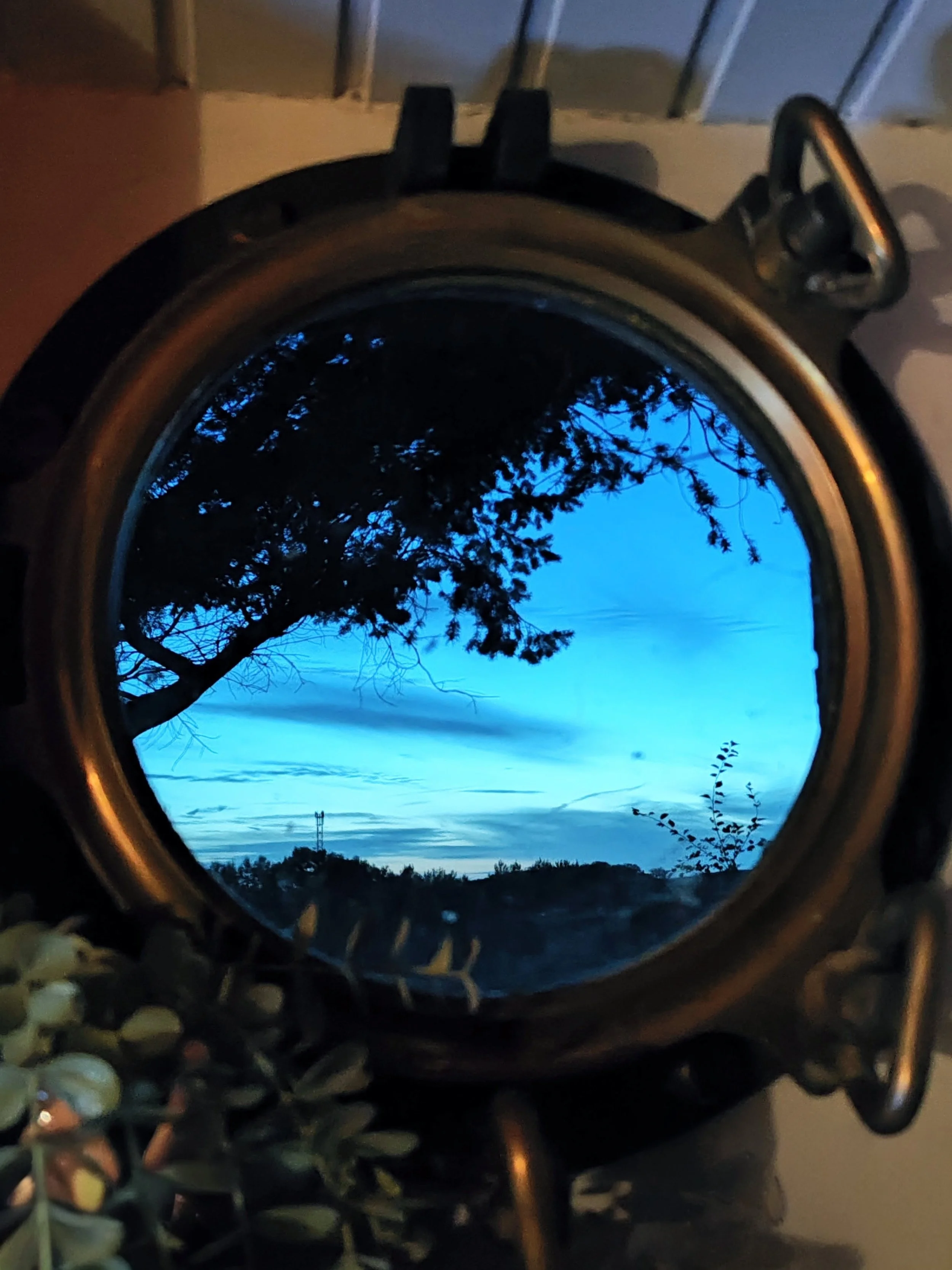 Der Blick durch ein runden, metallisches Fenster auf eine blaue Himmel mit Wolken und Silhouetten von Bäumen und einer Leitung im Hintergrund.
