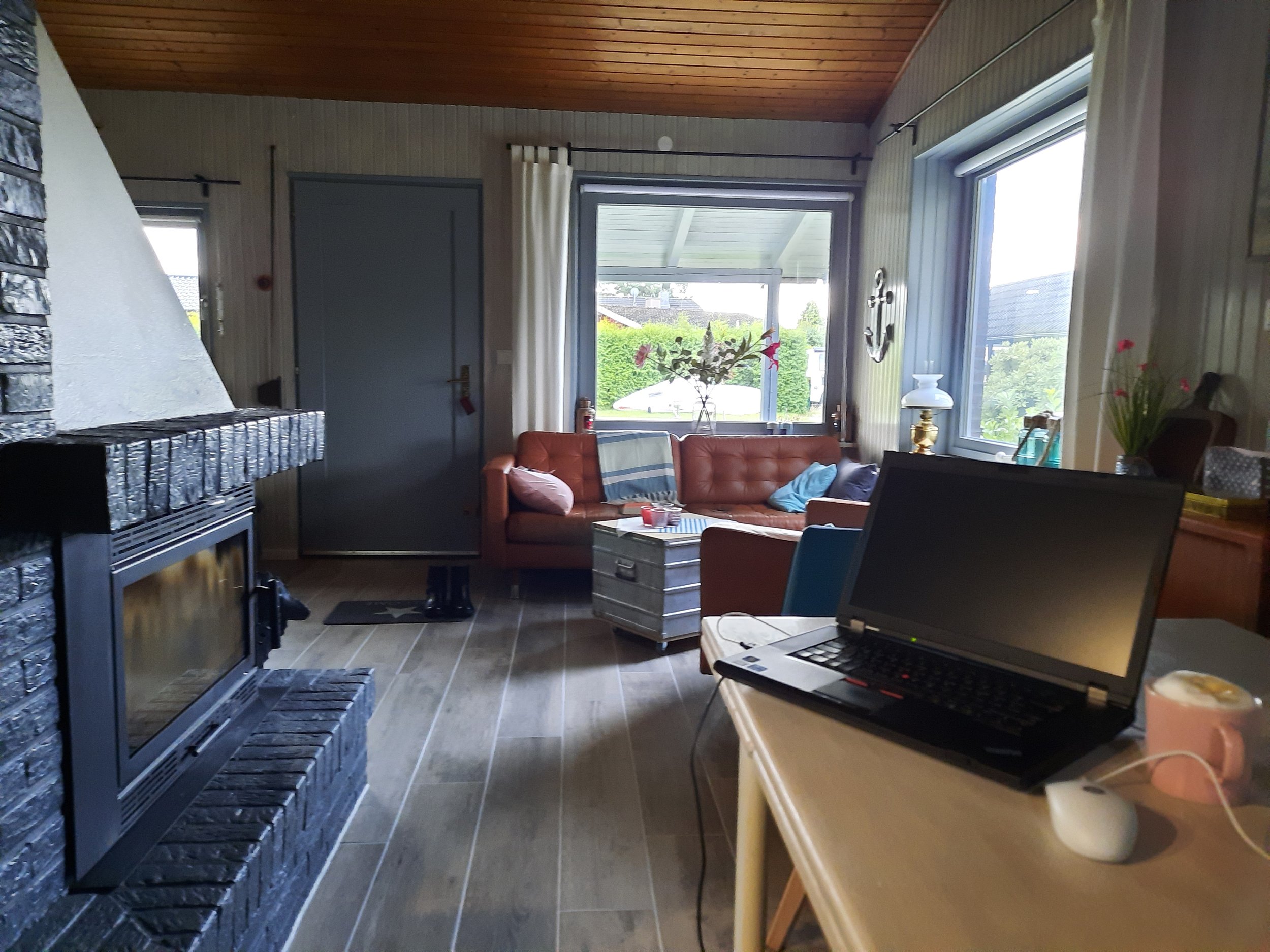 Gemütliches Wohnzimmer mit Kaminofen, Laptop auf Tisch, braunes Ledersofa, großes Fenster, Holzboden und Dekorationen.