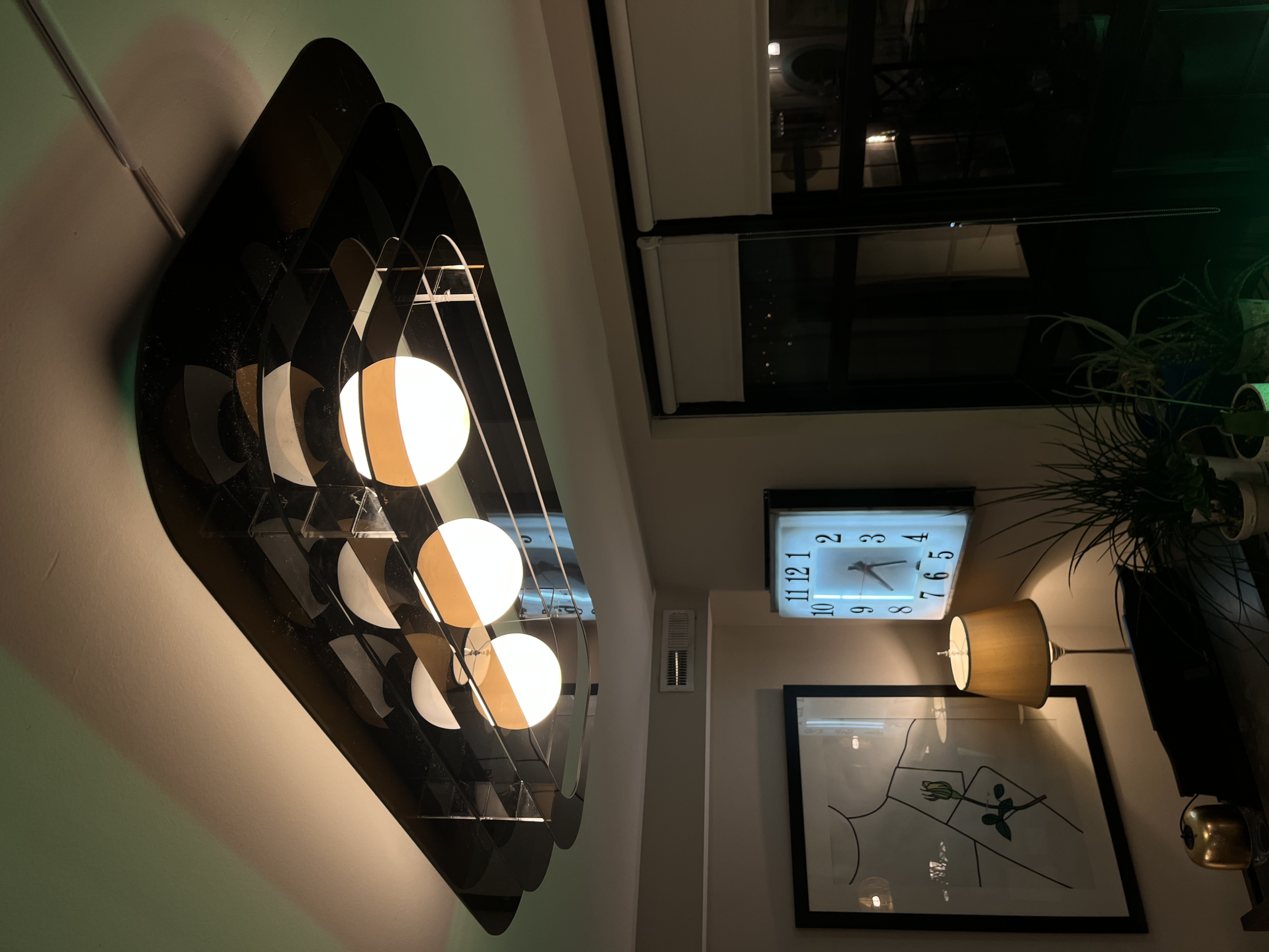 Indoor scene with a black acrylic light fixture holding three illuminated white spherical lights, a white clock with black numbers and hands, a tall corner plant, and a window showing a dark outdoor scene.