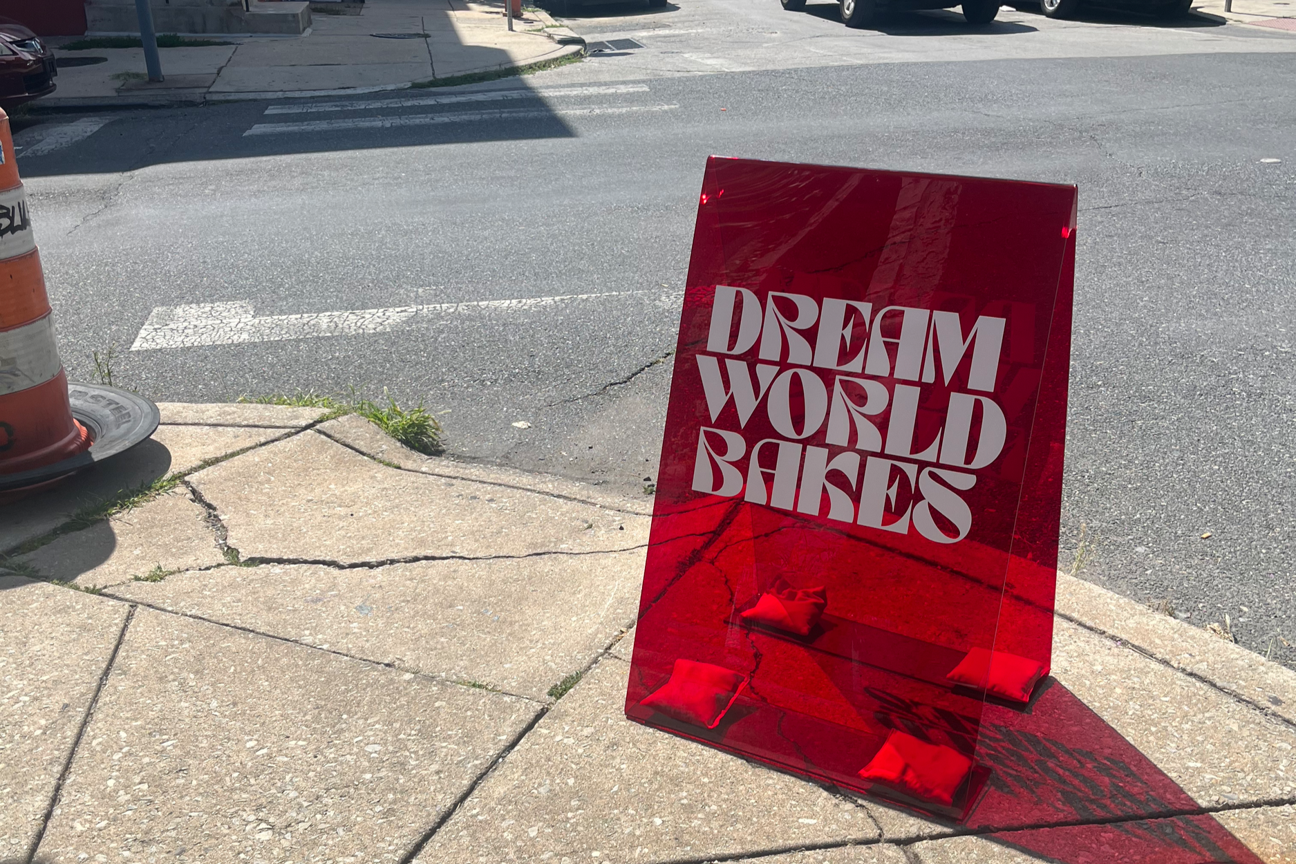 A red, translucent acrylic sign with the words "DREAM WORLD BAKES" in white, positioned on the sidewalk near the street, with cars parked in the background and a street curb.