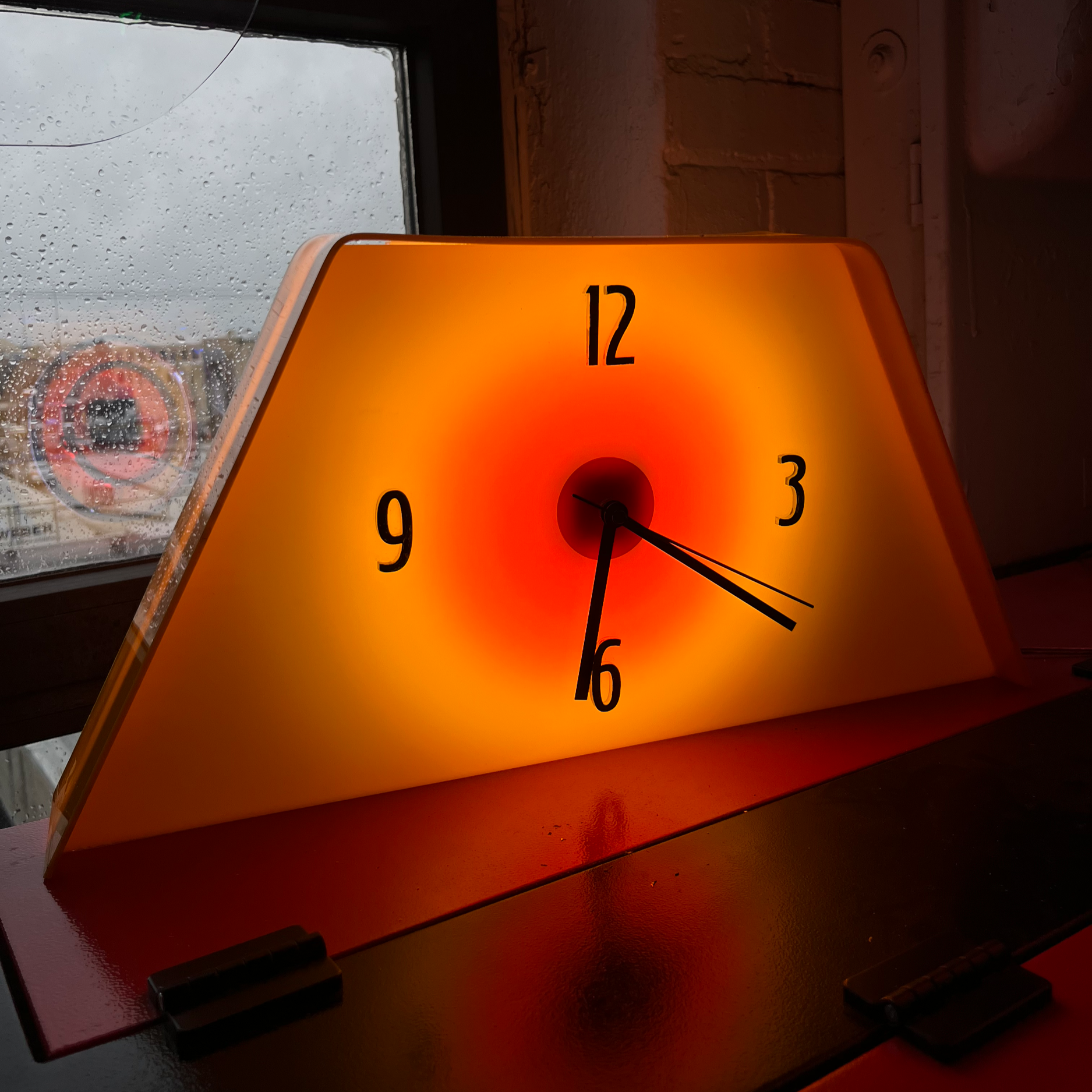 A back-lit orange clock with black numerals and black hands, displaying the time as approximately 5:30, is placed on a dark surface near a window with rain droplets, in a room with brick walls.