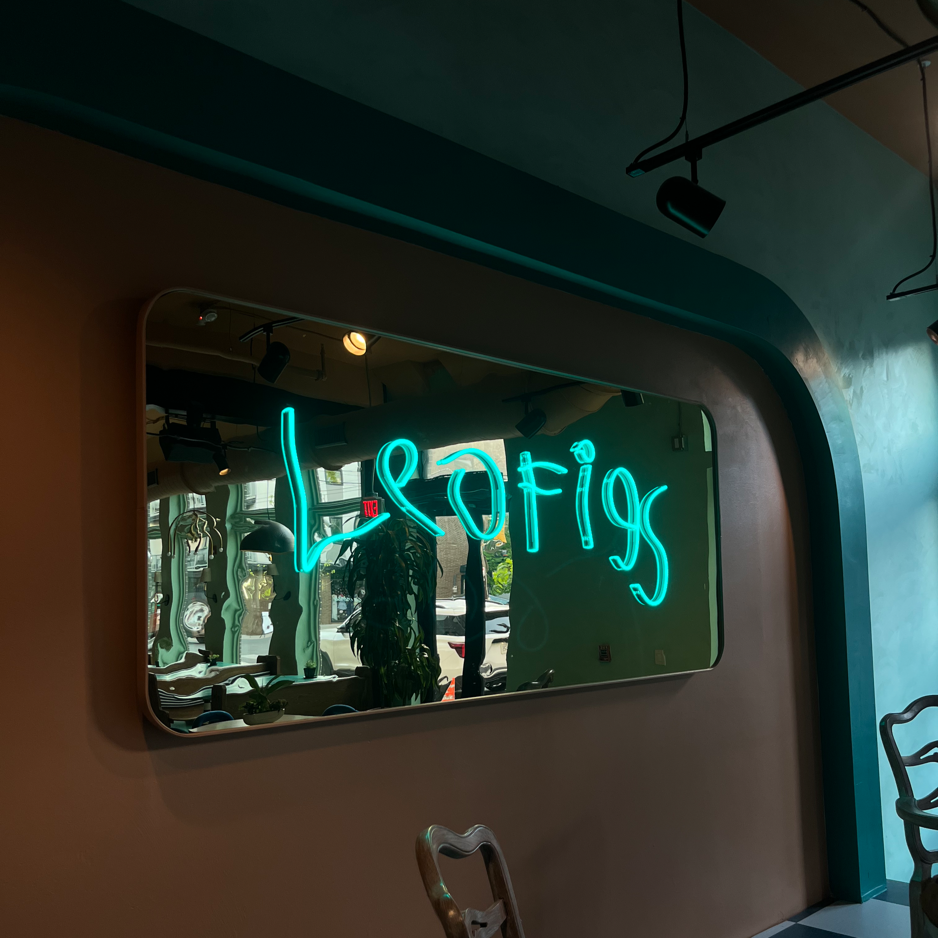 Interior view of a room with a large mirror on the wall, reflecting plants and hanging lights. A neon sign reads 'leofigs' in bright green. Through the window, parked cars and a street are visible outside.
