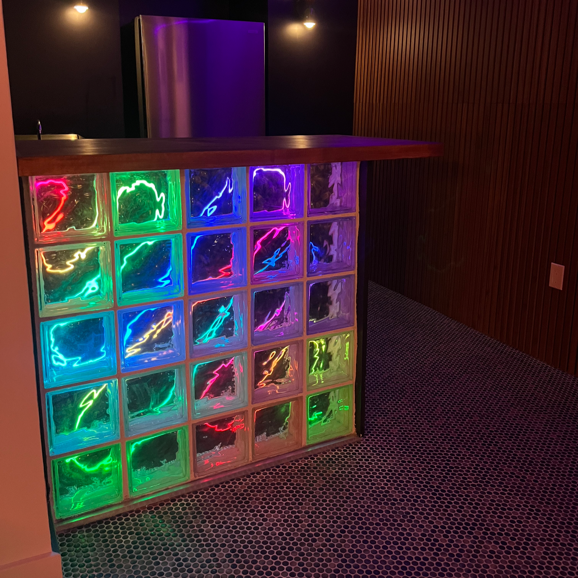 A dark room with a drink fridge illuminated by purple lights, a wooden bar counter, a wall with vertical wooden slats, and glass blocks with colorful neon lighting at the bottom.