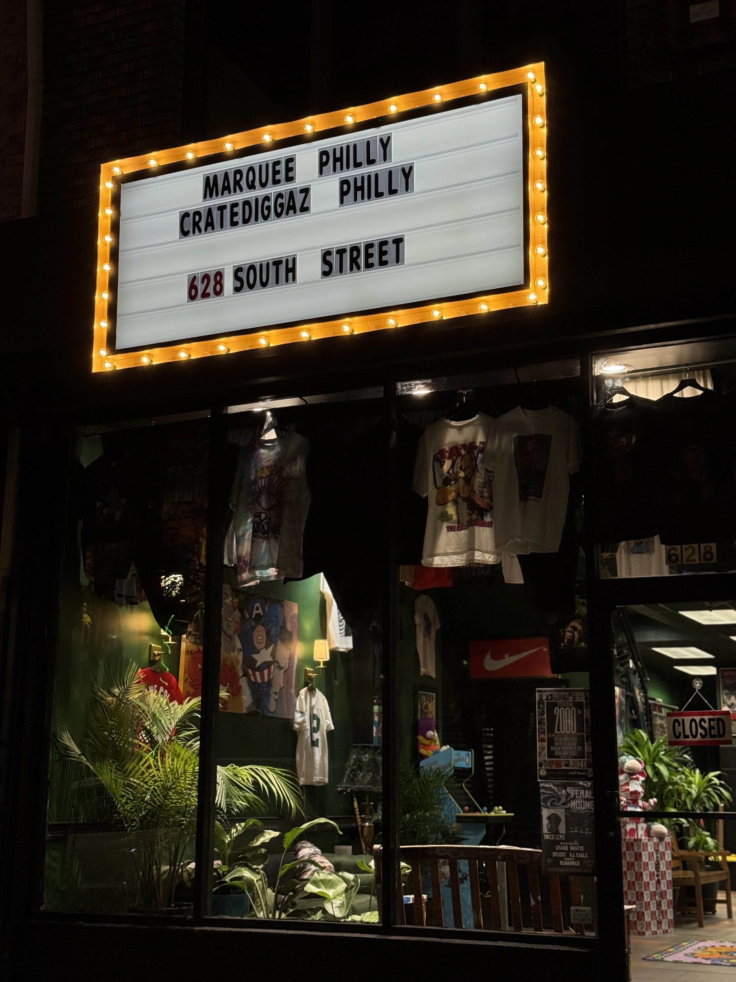 Nighttime view of a store window decorated with colorful posters, shirts, plants, and lighting. A marquee sign with bulbs displays text including 'Marquee Philly,' 'CrateDiggaz Philly,' and the address '628 South Street.' 
