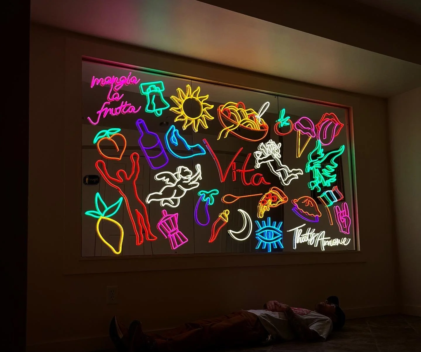 Colorful neon sign with various illustrations including a sun, bell, ice cream cone, pasta bowl, cherries, peach, bottle, angel, pizza slice, moon, eye, and text reading 'Vita' and 'That's Awesome', mounted on a wall. A person is lying on the floor i