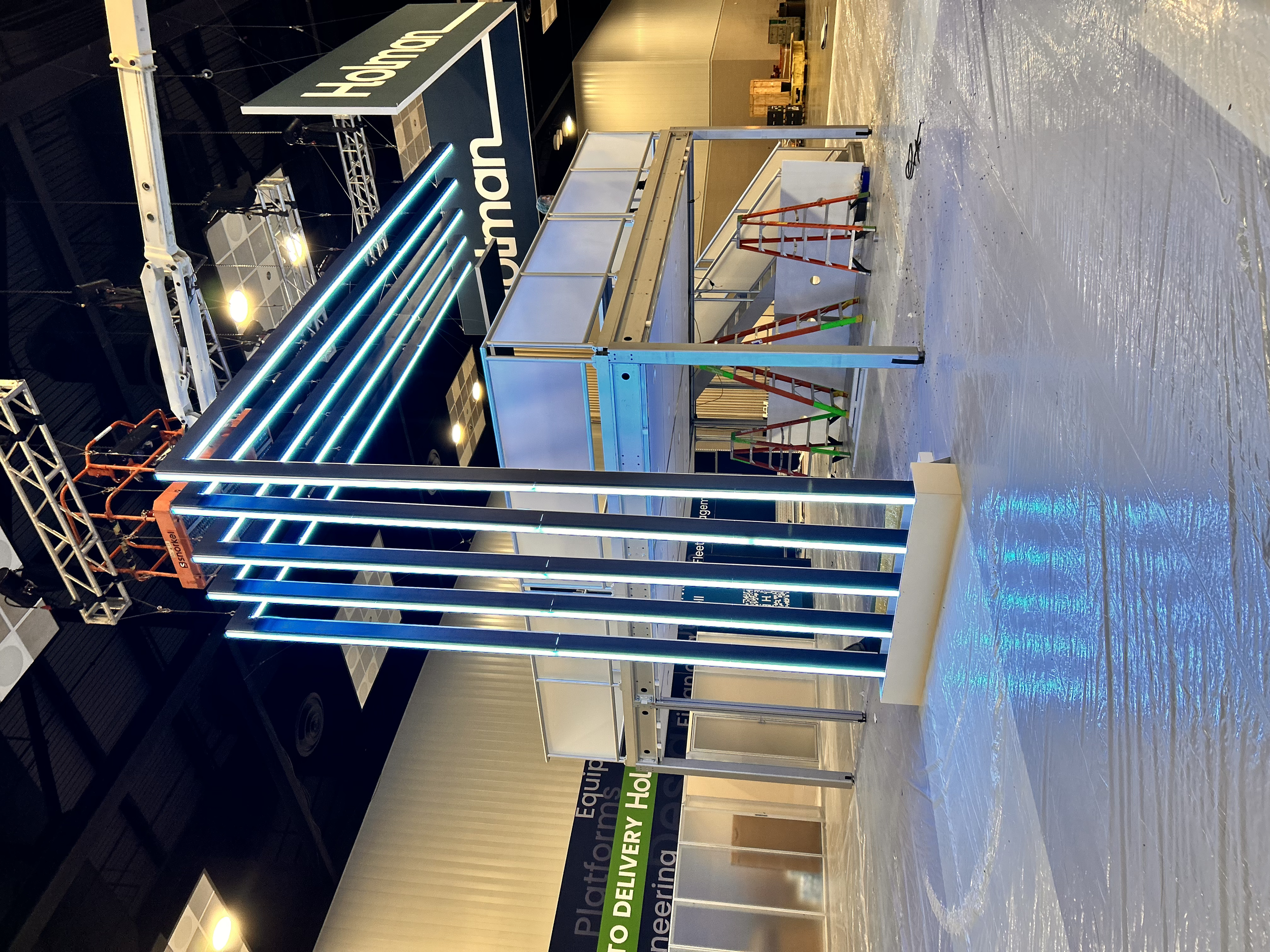 Empty retail kiosk with a metal frame, LED light strips, and a partially assembled sign. The background shows a large sign reading 'Holland' and a black ceiling with industrial lighting.