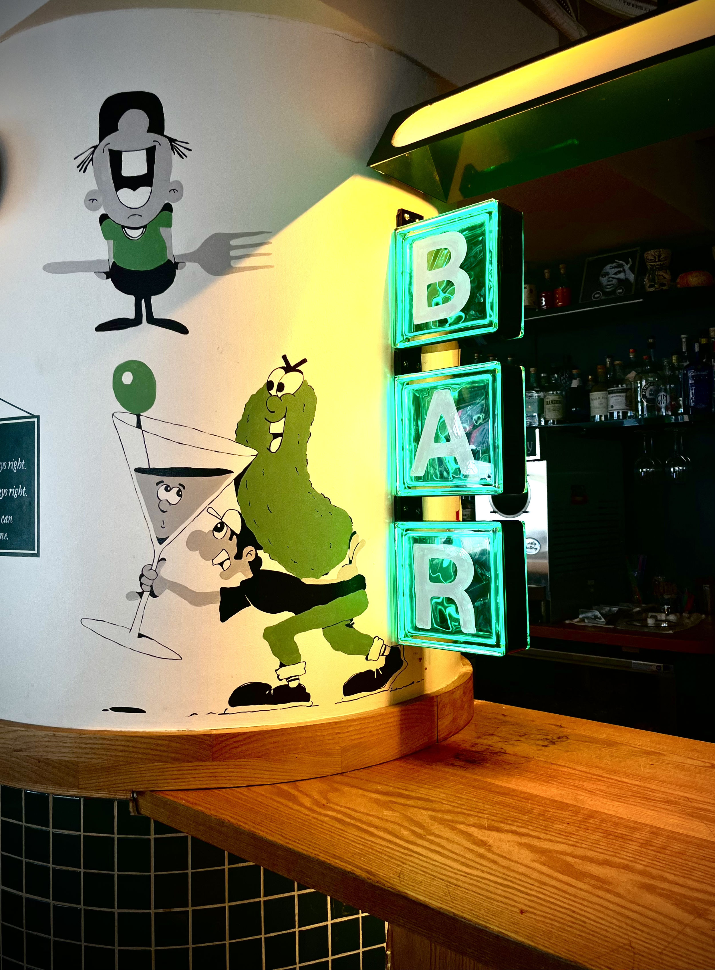 Colorful mural featuring cartoon characters including a woman with pigtails, a green creature, and a woman in a cocktail glass, next to illuminated glass bricks spelling "BAR."
