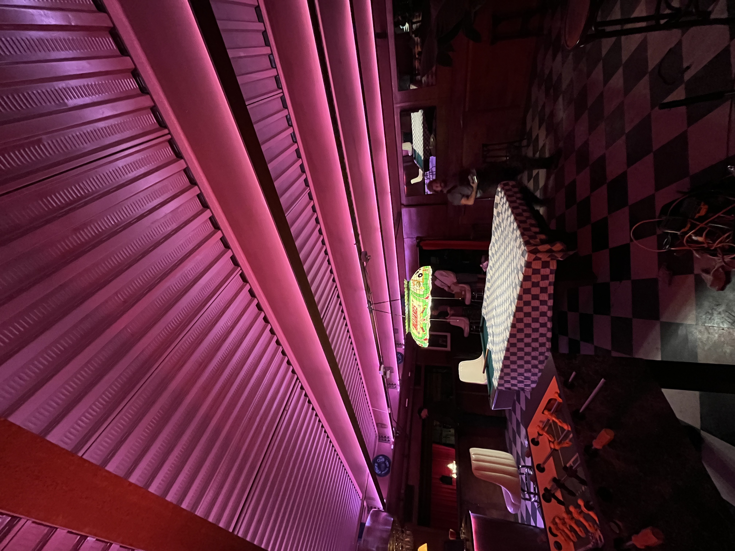 A dimly lit restaurant or bar interior with purple-pink neon lighting, checkered tablecloths, and a woman standing at the counter.