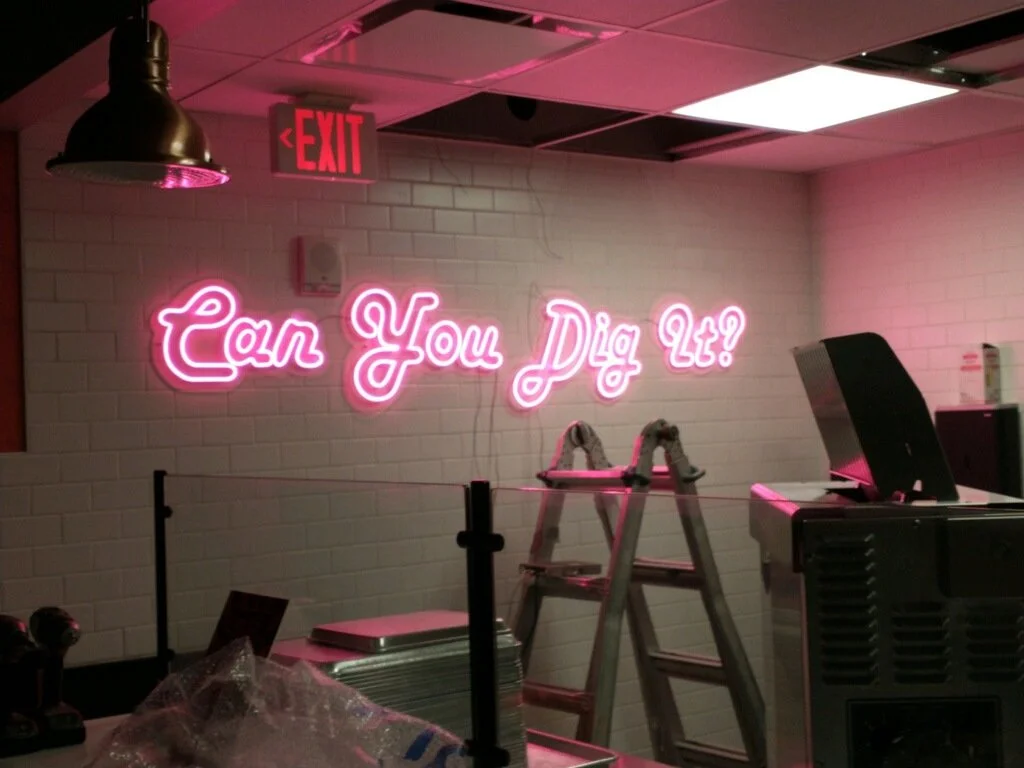 Neon pink sign on a white brick wall reads, 'Can You Dig It?'. 
