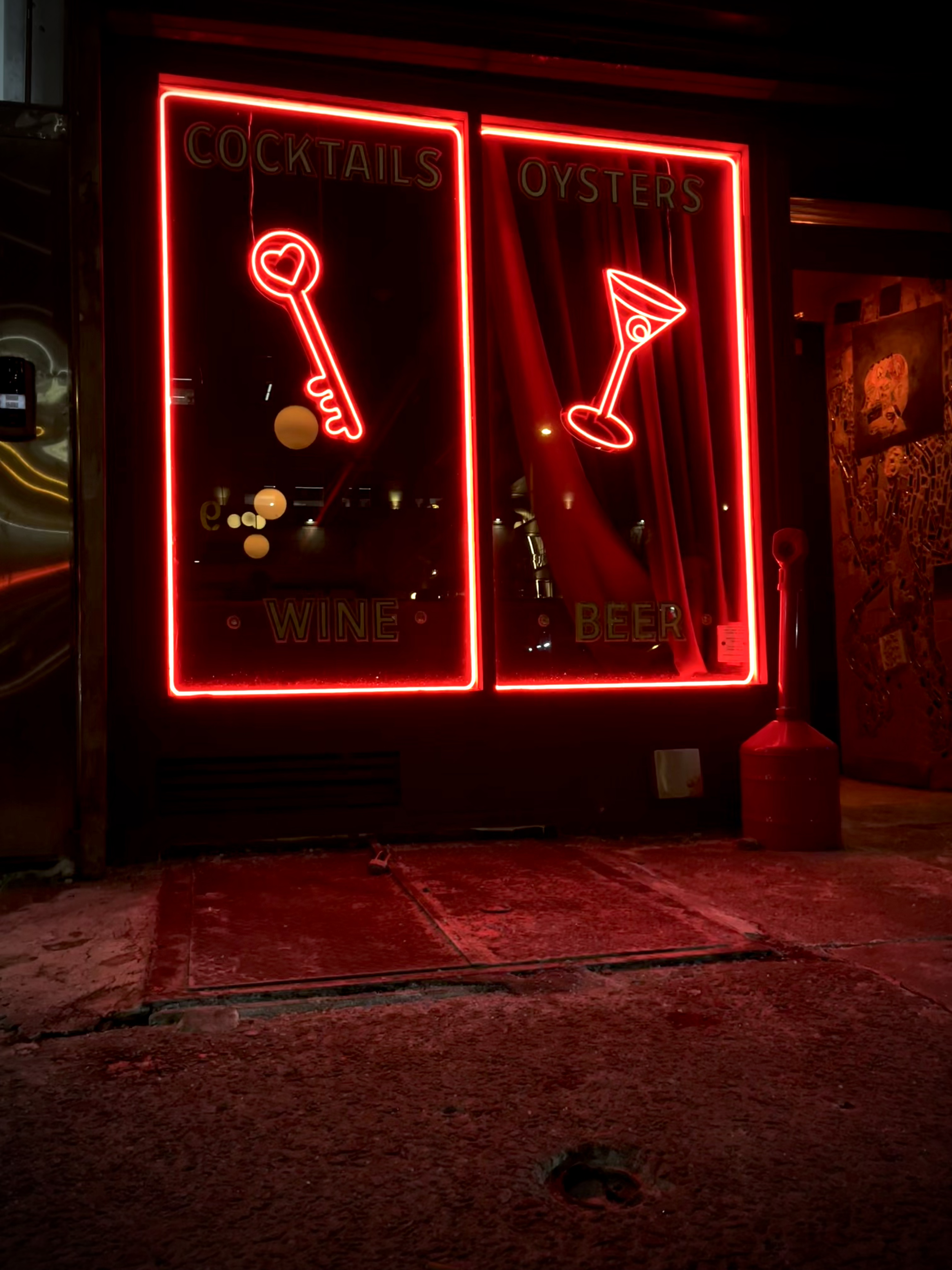Neon signs advertising cocktails, oysters, wine, beer, and a cocktail glass, illuminated in red on a dark background outside a bar or restaurant.