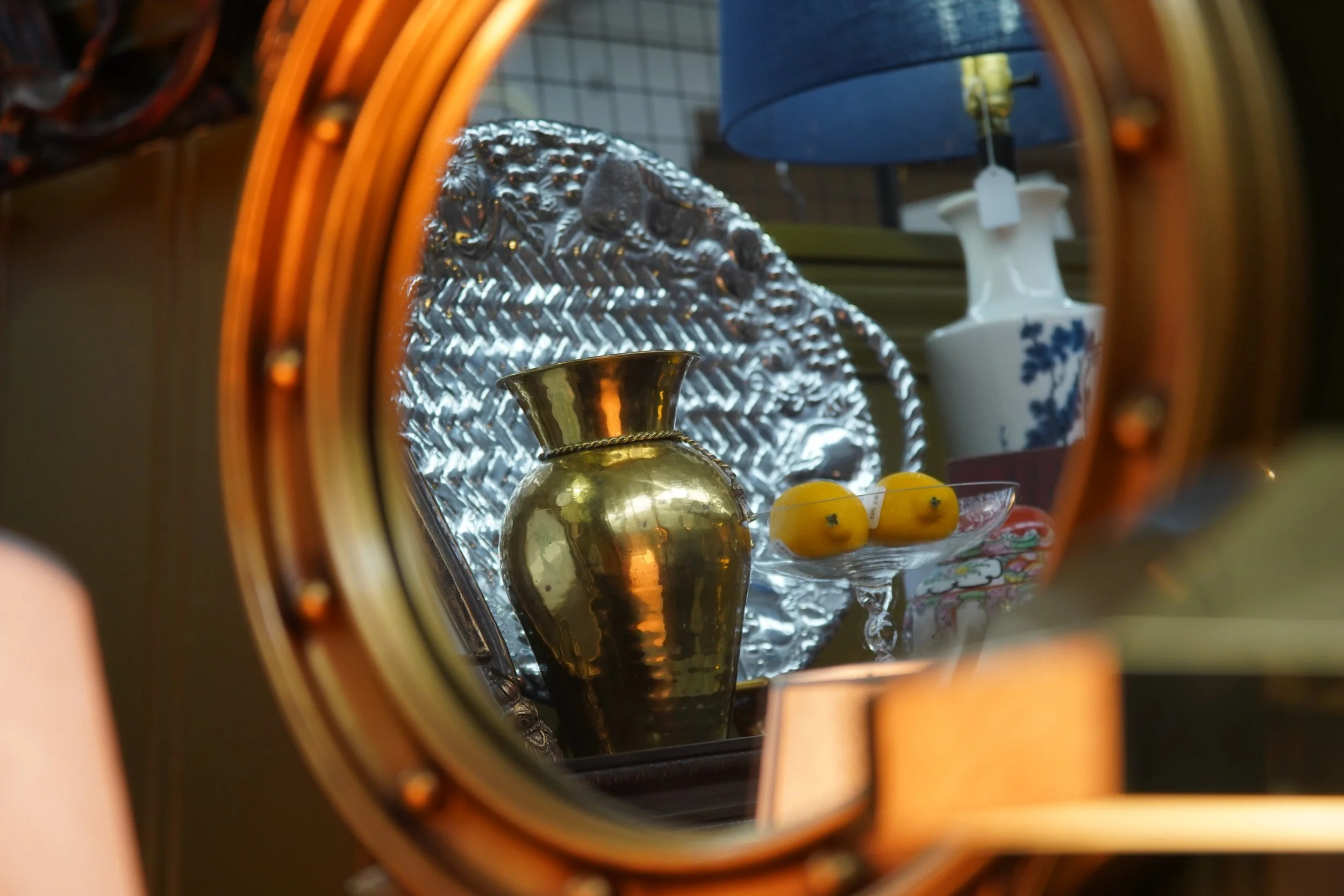 Reflection of a decorative display in a mirror showing a brass vase, a glass dish with yellow fruit, a white vase with blue floral pattern, and a dark blue lampshade.