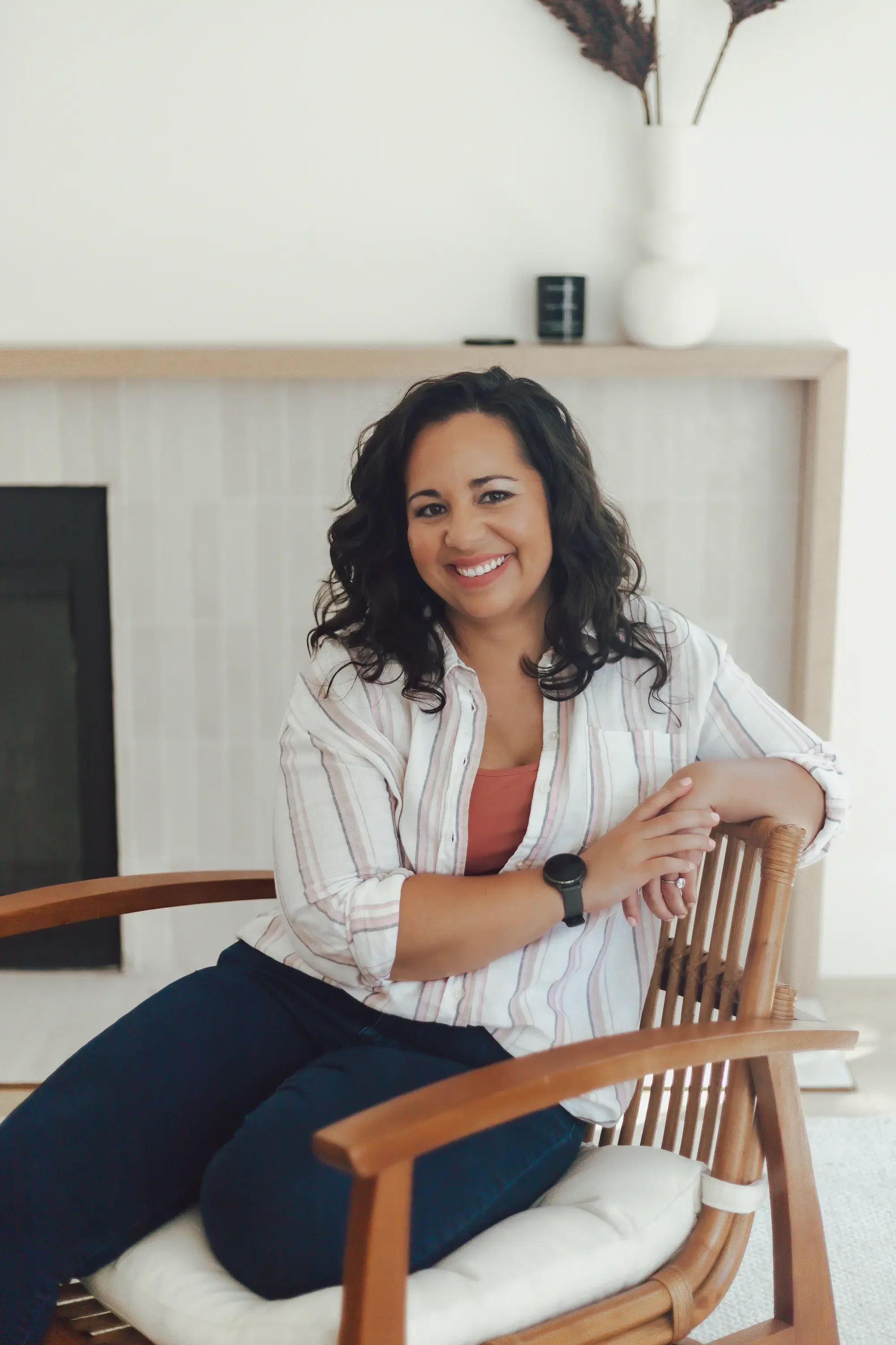 Gottman couples therapist with dark long curly hair smiling as she sits in a wooden chair with a white fireplace in the background of her therapy office.