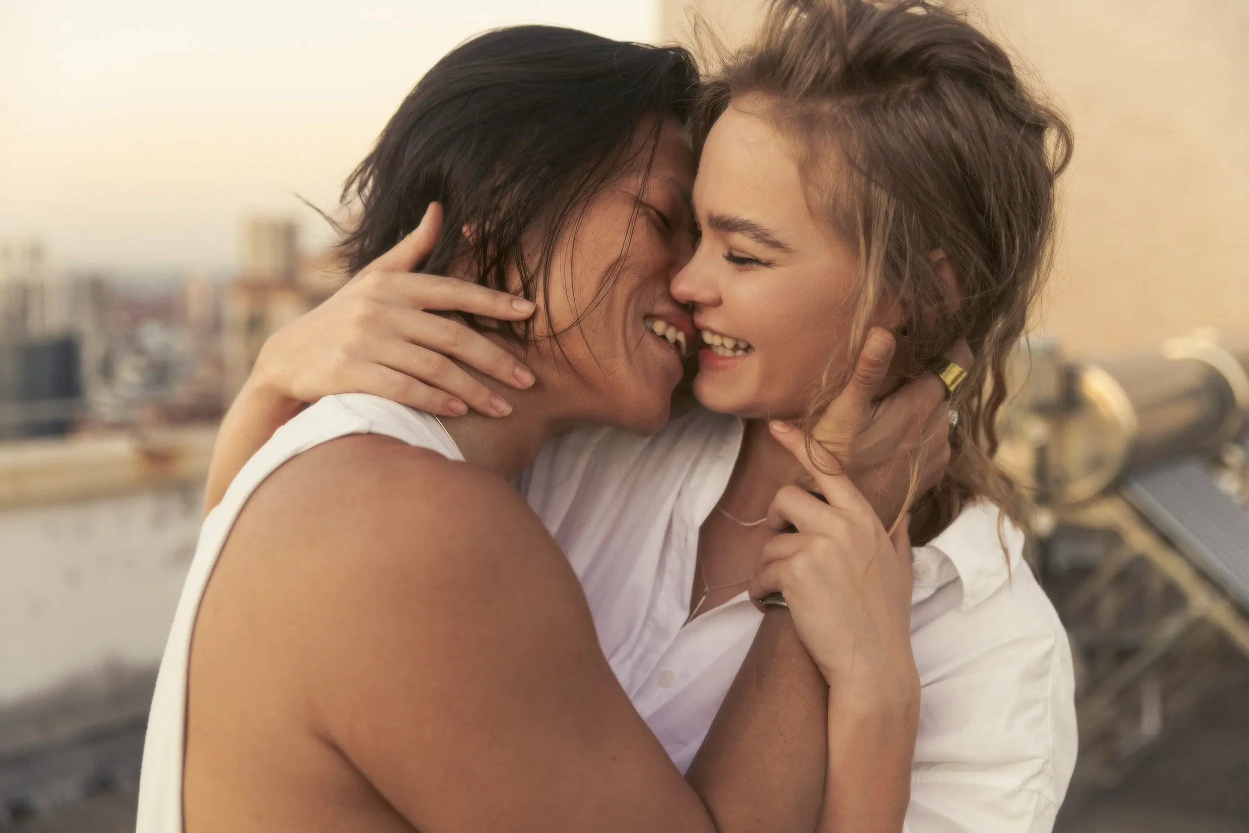 A couples is embracing, touching foreheads and smiling, with a city skyline in the background during sunset.