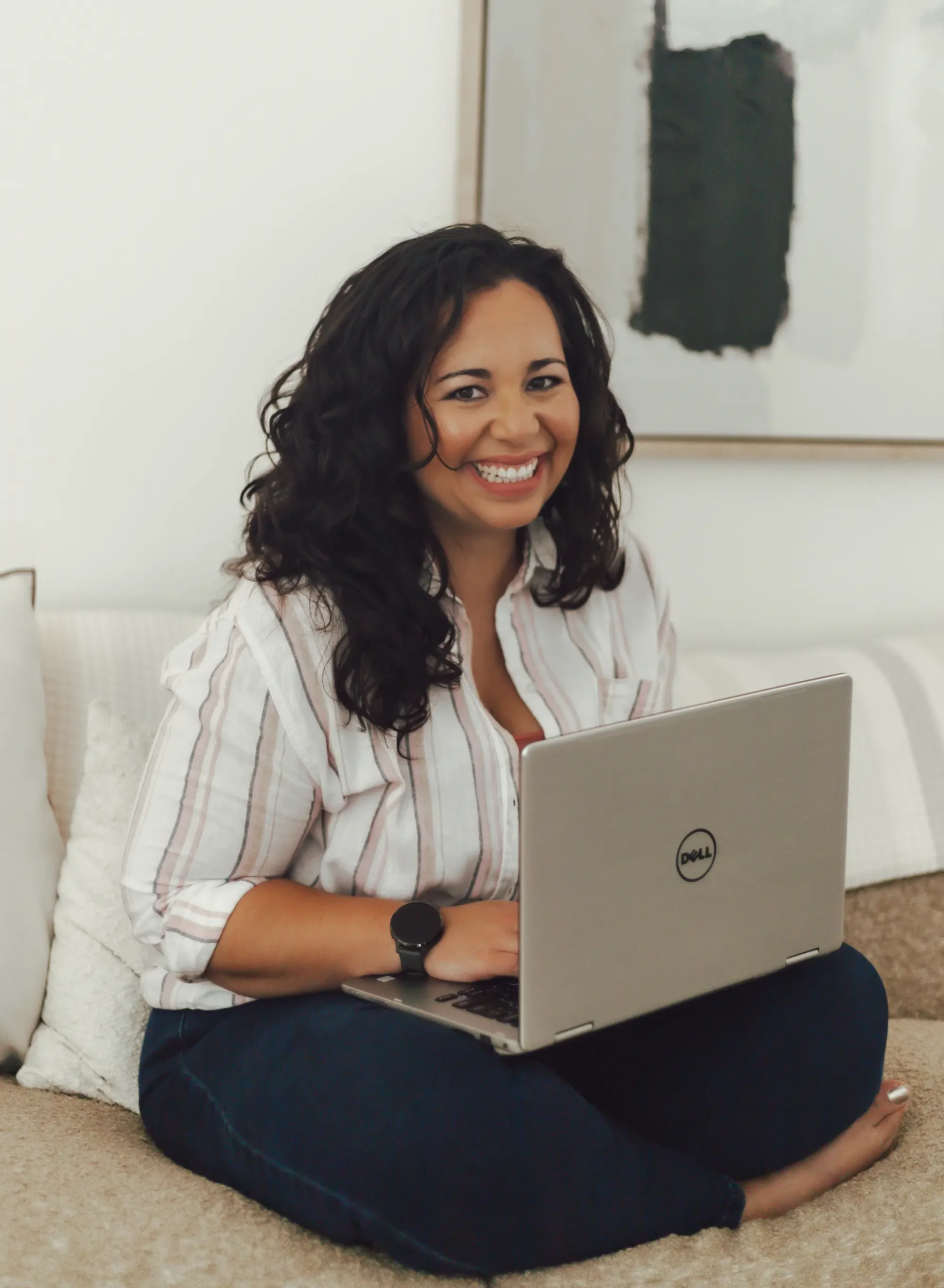 Woman with long brown curly hair and white striped button up smiling as she holds her silver laptop while sitting on a tan couch.