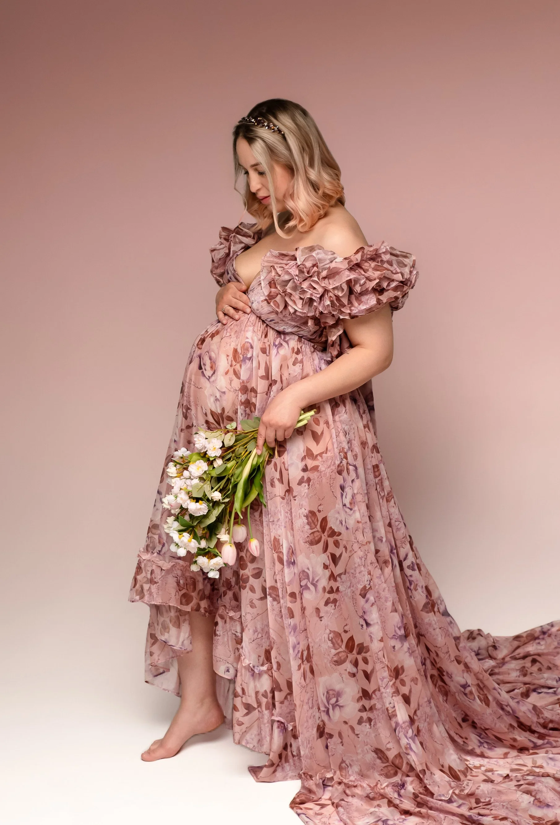 A pregnant woman wearing a pink floral organza gown and holding a flower bouquet, in a maternity photosession in Calgary