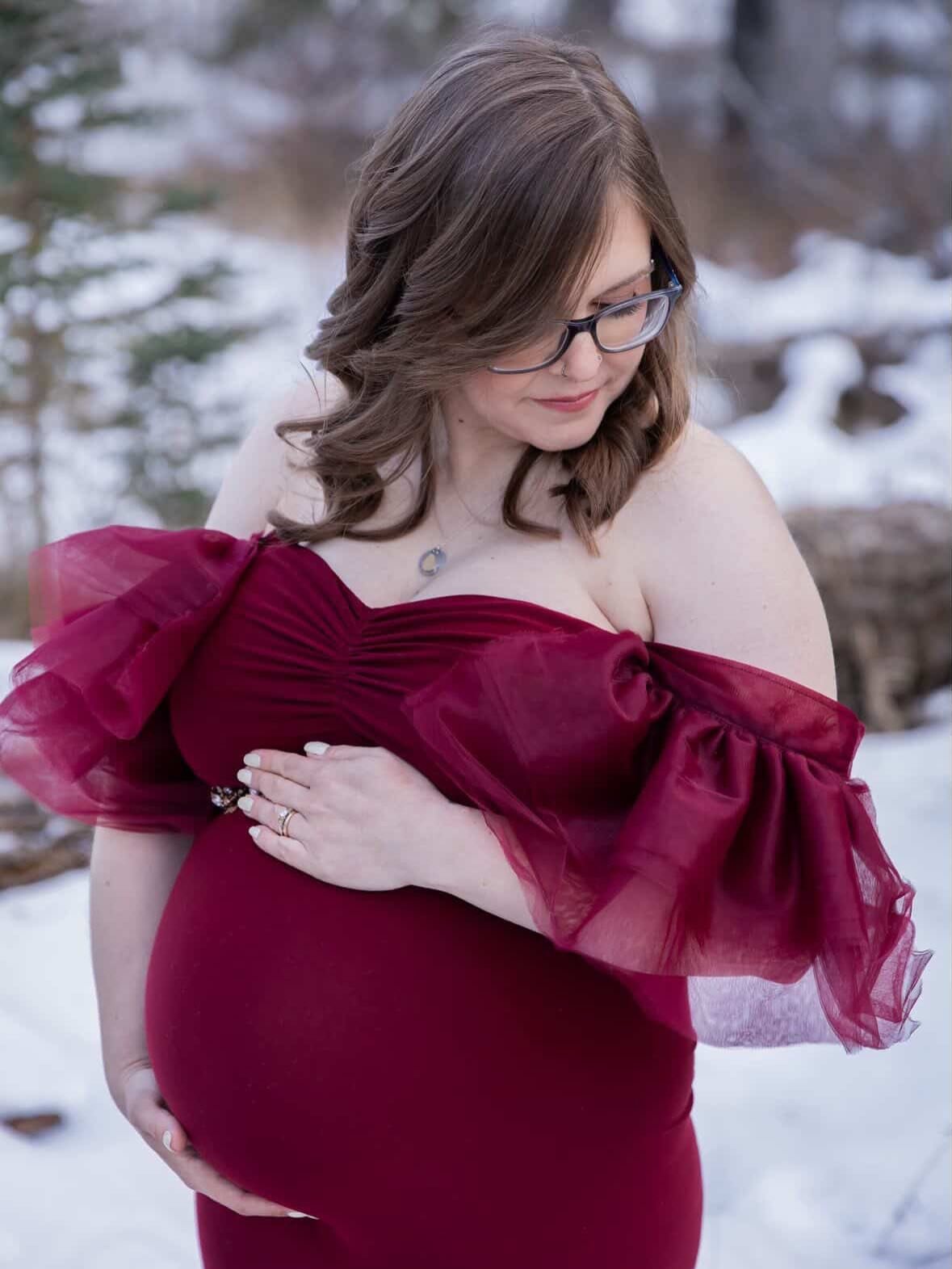 A pregnant woman wearing a red bodycon gown with ruffled off-the-shoulder tulle sleeves and long tulle train, in outdoor winter photoshoot
