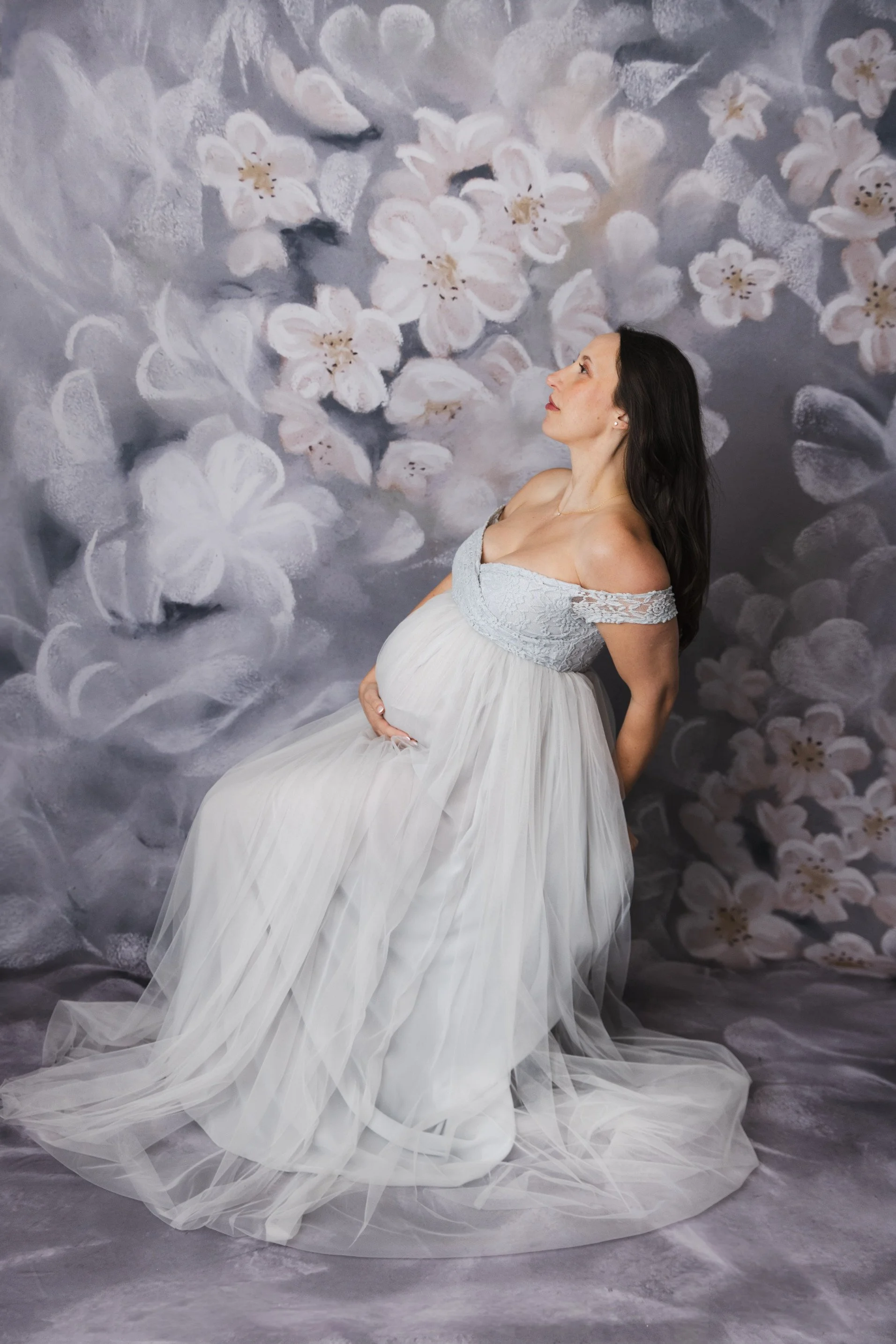 A pregnant woman wearing a silver tulle rental dress with off-the-shoulder embroidered corset, in a photoshoot with a floral backdrop
