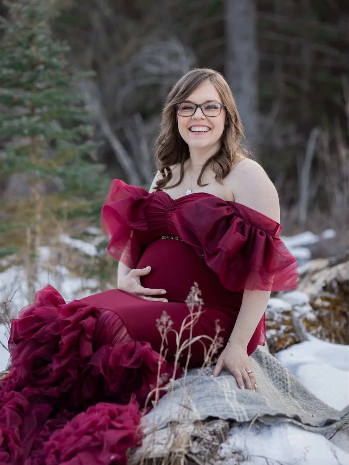 A pregnant woman wearing a red bodycon gown with ruffled off-the-shoulder tulle sleeves and long tulle train, in outdoor winter photoshoot