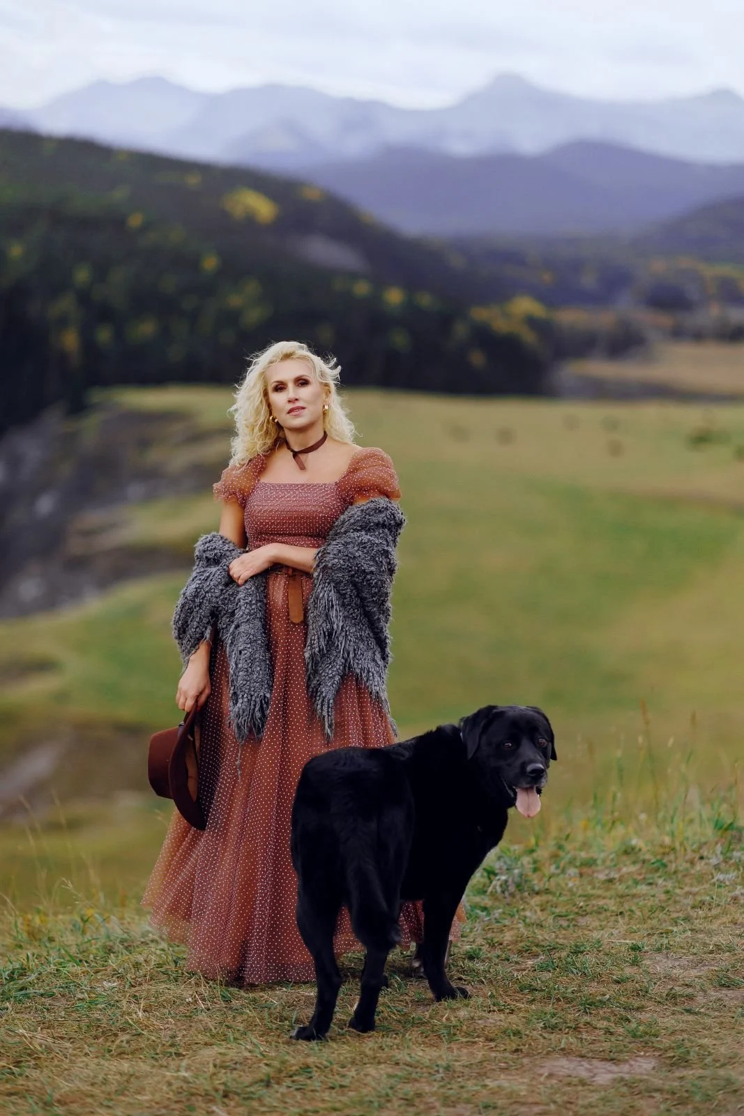A woman wearing a rust brown tulle rental gown with white polkadots, standing with a black dog in outdoor photoshoot