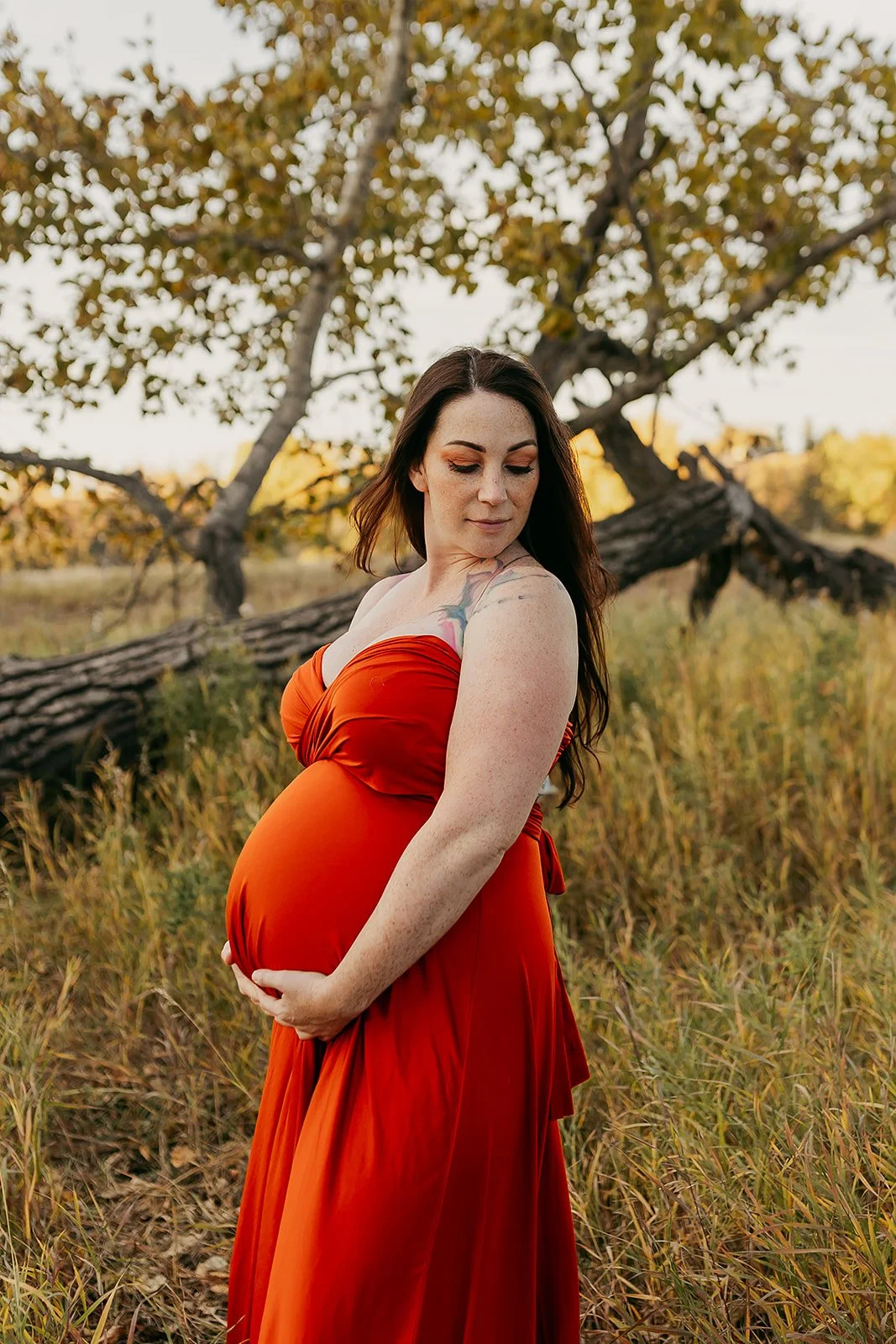 A pregnant woman wearing an orange red maternity infinity wrap dress in an autumn field outdoor photosession