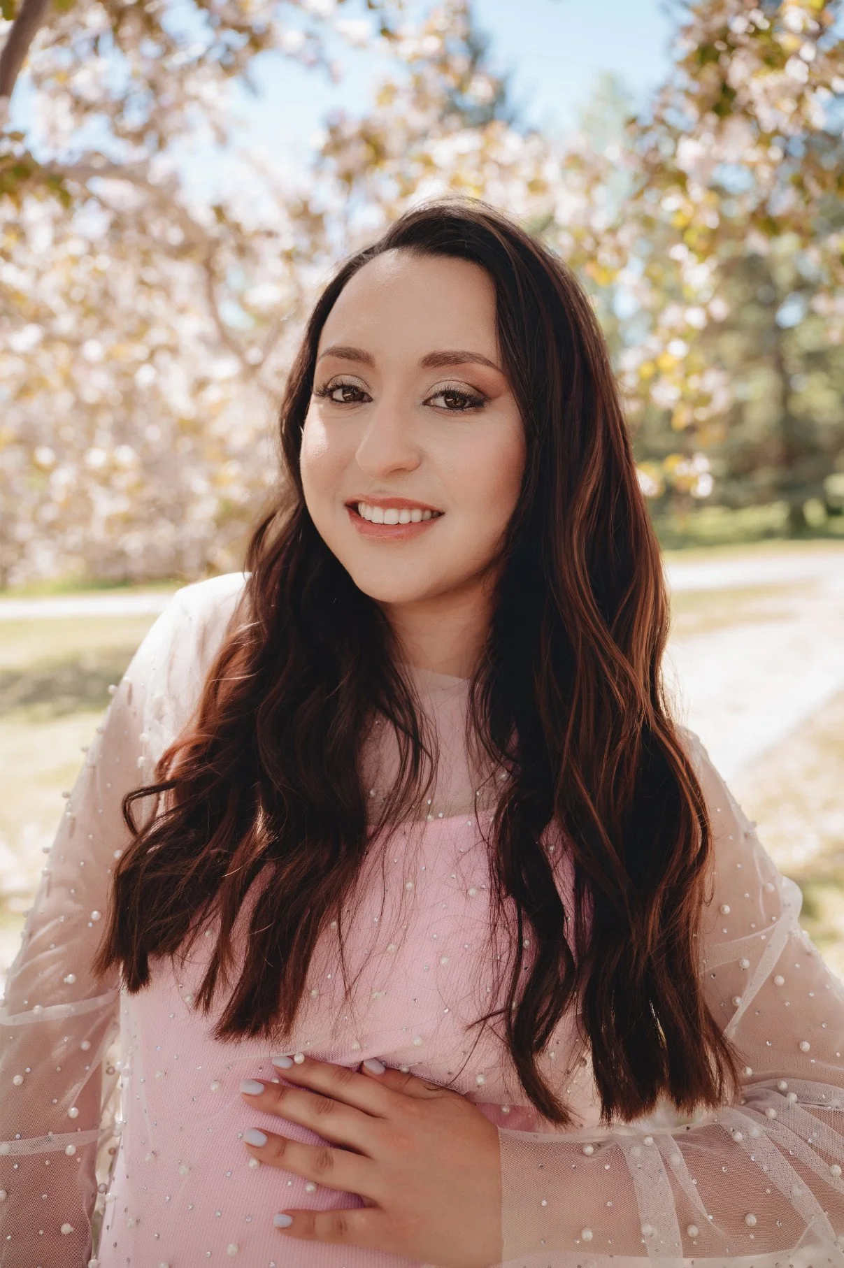 A pregnant woman wearing a cotton candy pink fitted dress with sheer layer of pearl embellishments throughout the dress, and long sheer sleeves, in outdoor spring maternity photoshoot