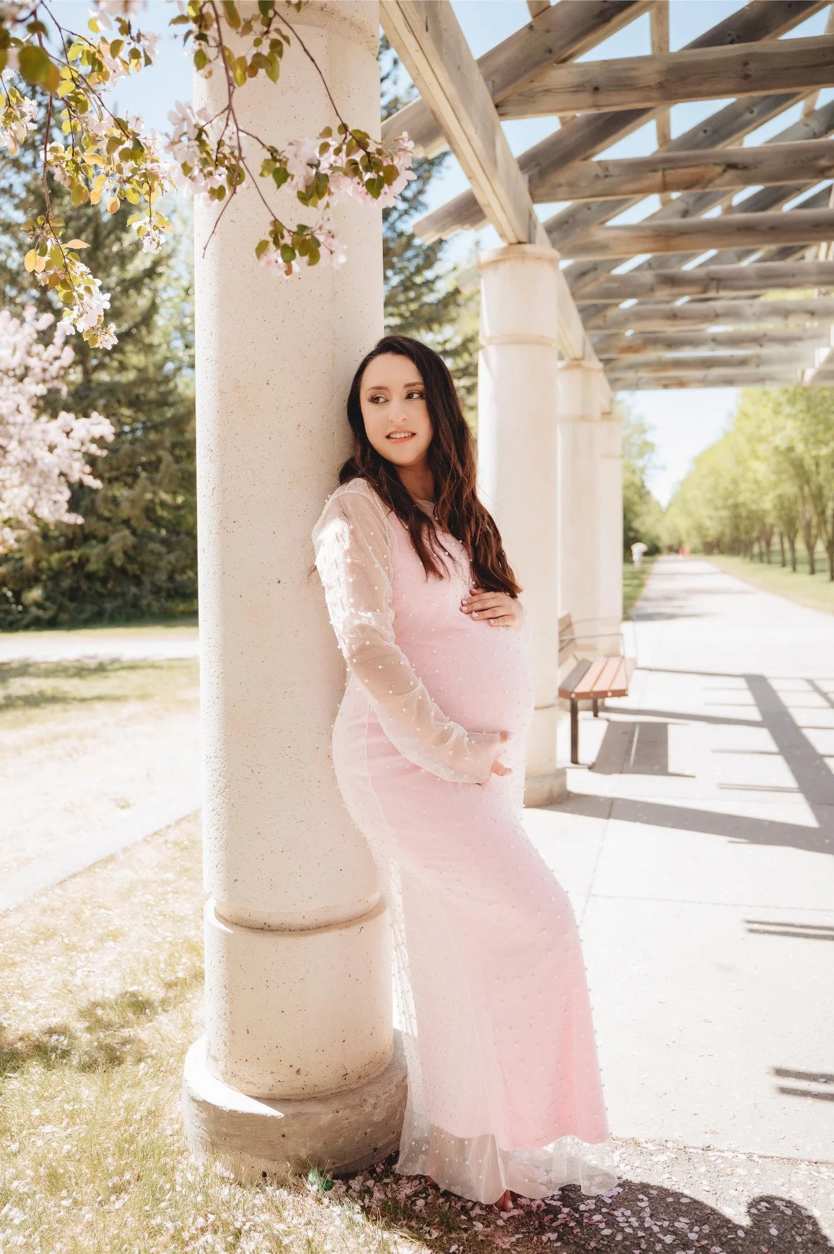 A pregnant woman wearing a cotton candy pink fitted dress with sheer layer of pearl embellishments throughout the dress, and long sheer sleeves, in outdoor spring maternity photoshoot