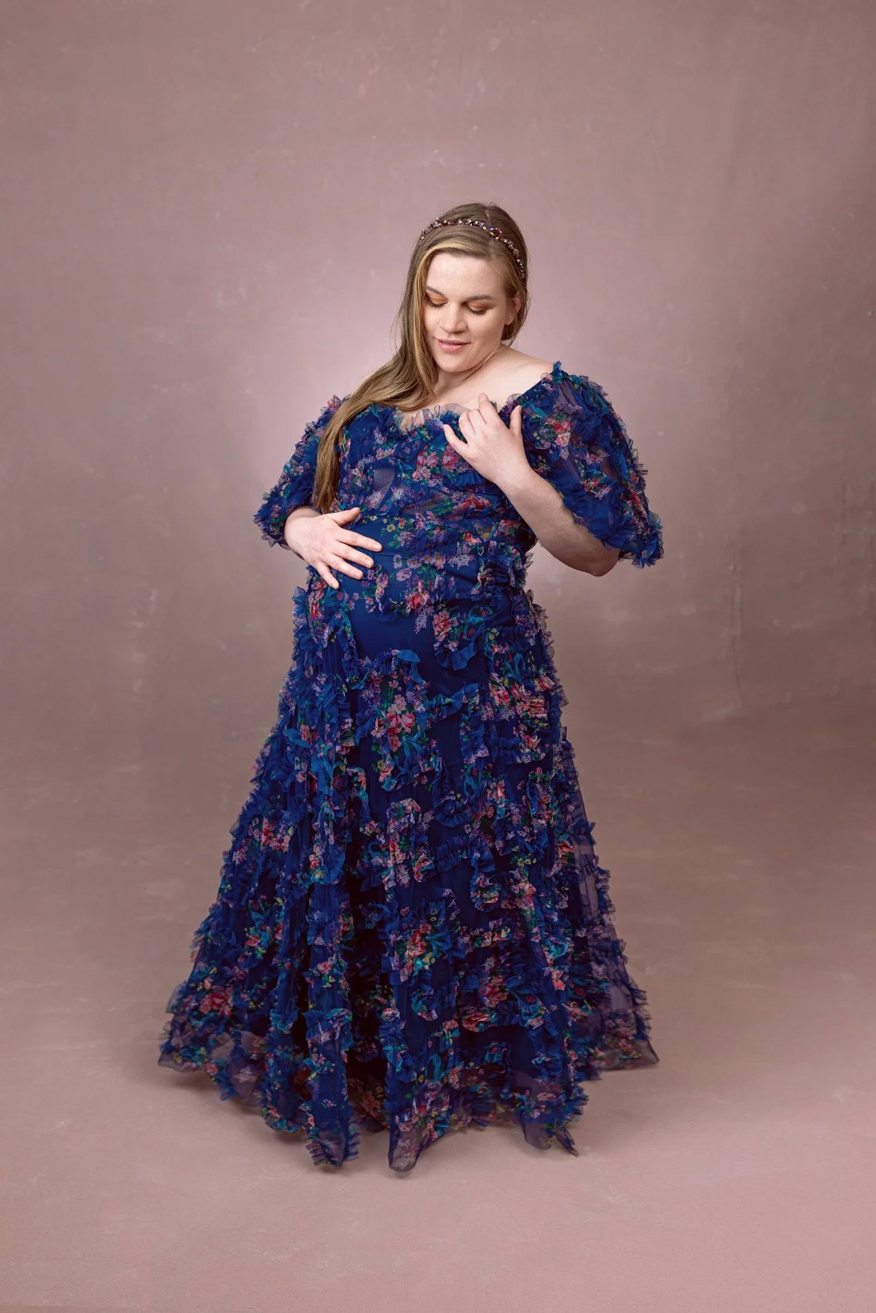 Pregnant woman wearing a navy blue dress with floral patterns and a lot of tulle texture, in an indoor studio maternity photoshoot setting