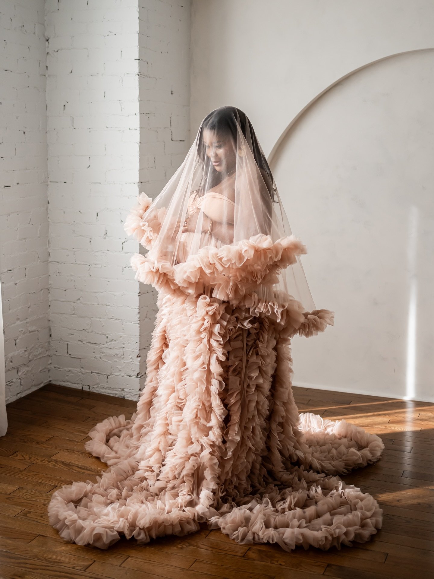 A pregnant woman wearing a ruffled tulle dusty rose gown, with sheer tulle veil over her head, in Calgary maternity studio session