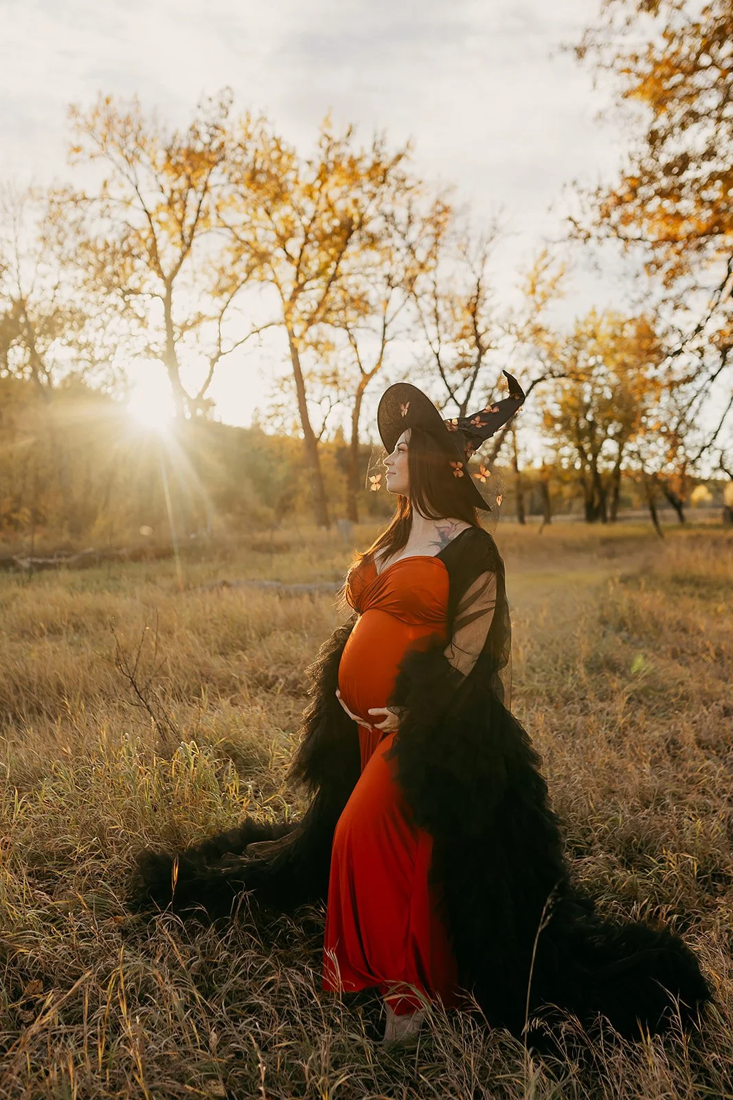 A pregnant woman wearing an orange infinity dress with a black tulle robe and witch hat in outdoor autumn maternity photosession