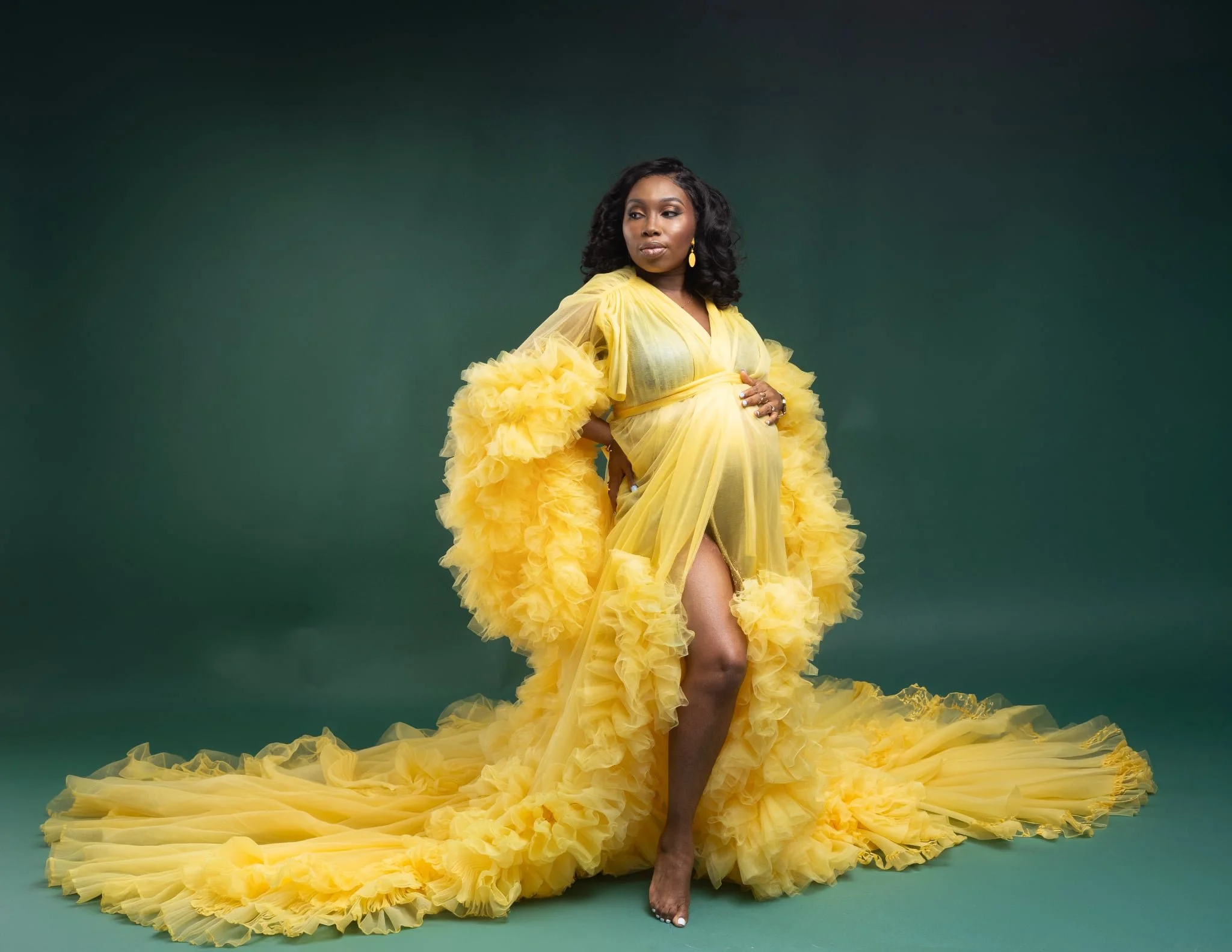A pregnant woman wearing a golden yellow sheer tulle maternity robe with open belly front, posing in a studio pregnancy photoshoot