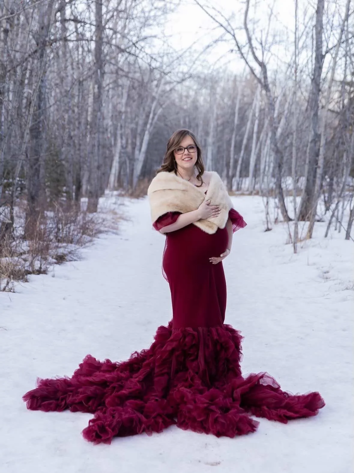 A pregnant woman wearing a red bodycon gown with ruffled off-the-shoulder tulle sleeves and long tulle train, in outdoor winter photoshoot