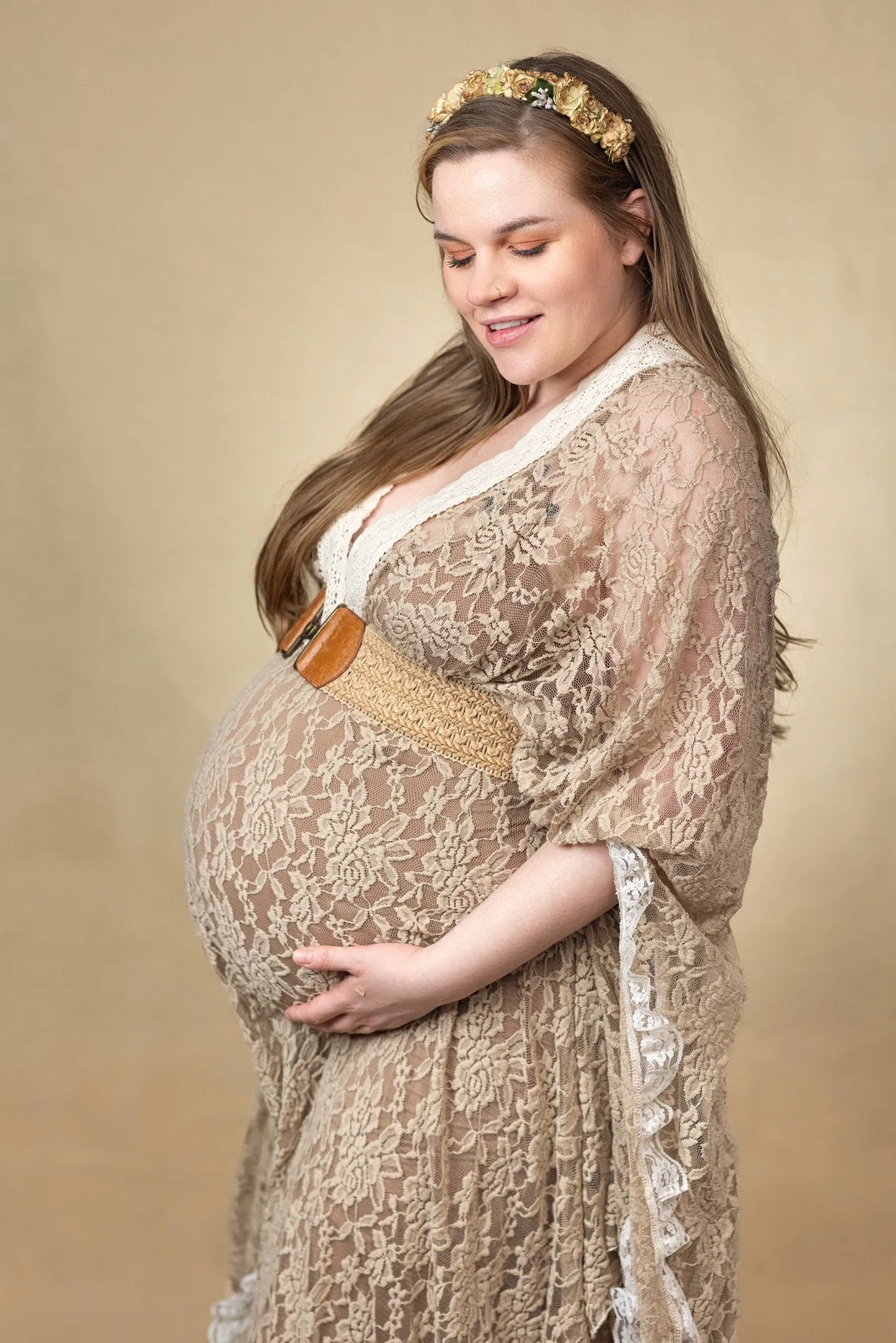 Close up of a pregnant woman, showing her wearing a lace khaki-coloured dress in an indoor photoshoot setting