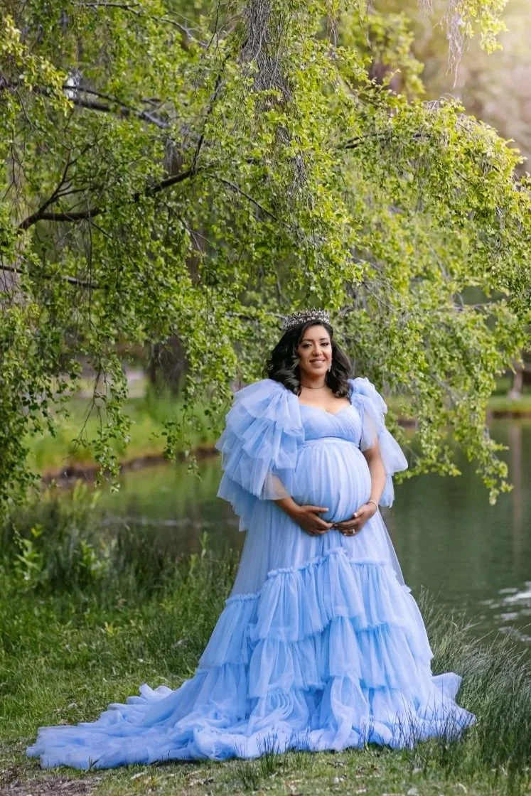 A pregnant woman wearing a baby blue tulle gown with puffy shoulders and long skirt in summer outdoor maternity photoshoot