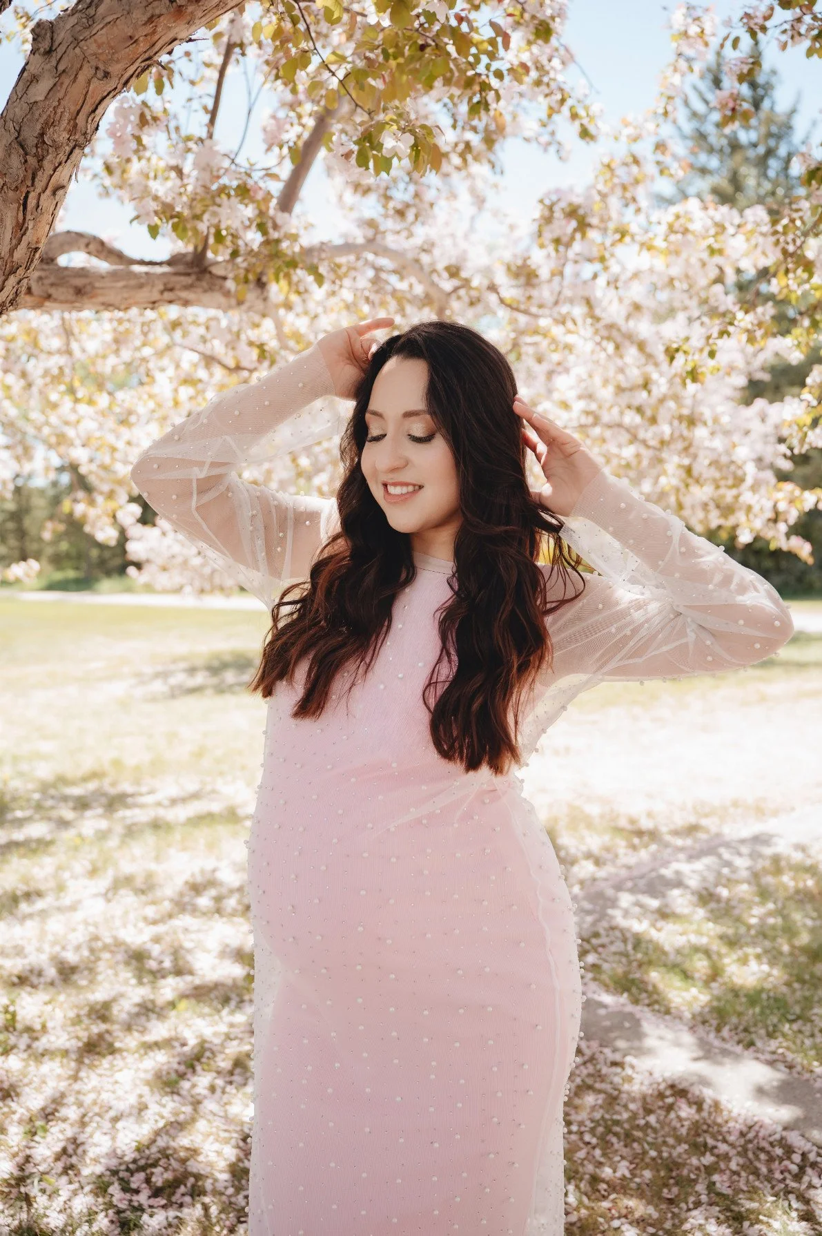 A pregnant woman wearing a cotton candy pink fitted dress with sheer layer of pearl embellishments throughout the dress, and long sheer sleeves, in outdoor spring maternity photoshoot