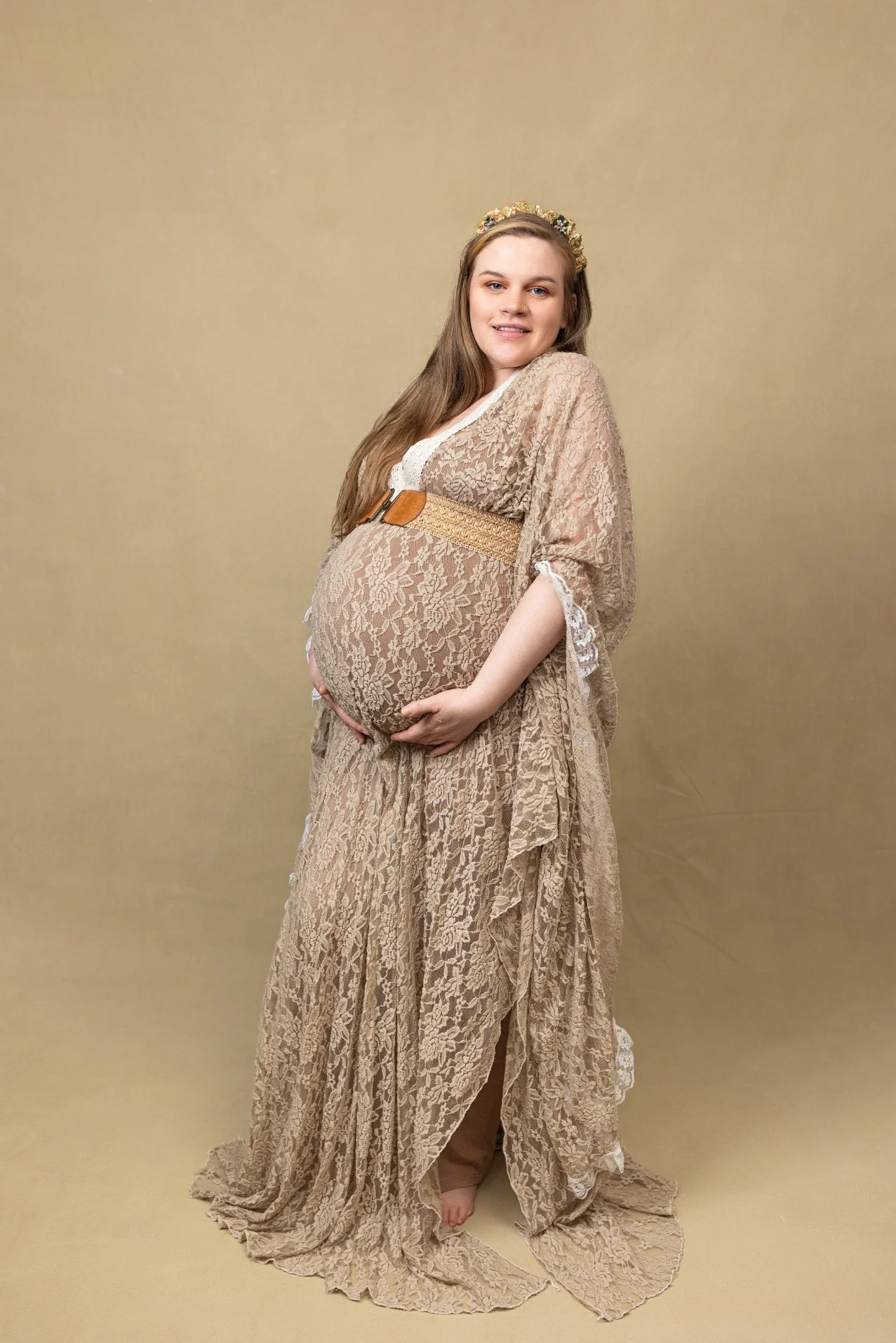 A pregnant woman wearing a khaki lace maternity dress, in an indoor photoshoot