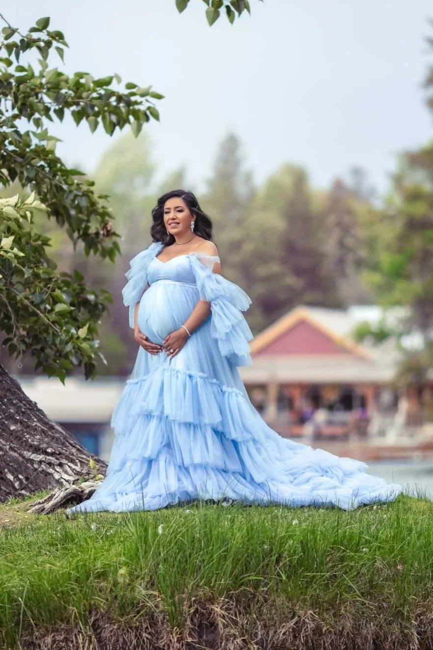A pregnant woman wearing a baby blue tulle gown with puffy shoulders and long skirt in summer outdoor maternity photoshoot