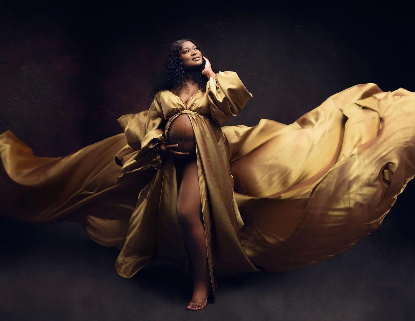 A pregnant woman wearing a flowing golden robe with a cutout in the middle, in a studio maternity photoshoot setting