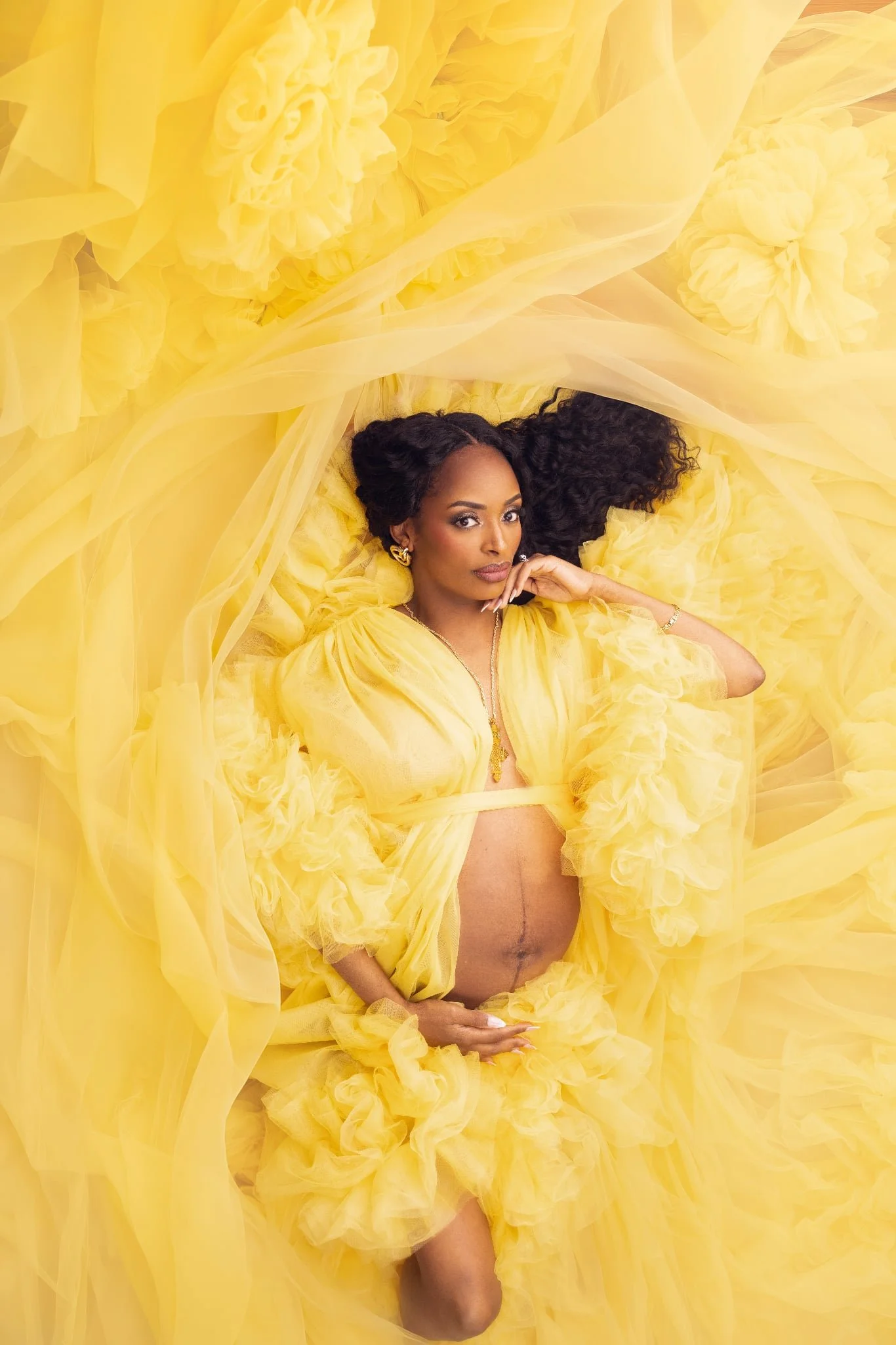 A pregnant woman enveloped in a golden yellow sheer tulle maternity robe, posing in a studio pregnancy photoshoot